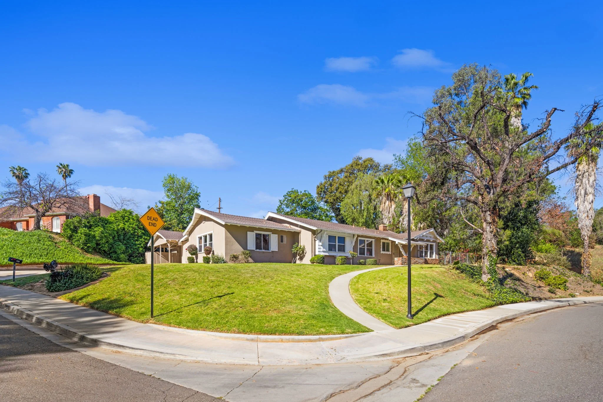 Residential neighborhood with a house, trees, a lamp post, a sidewalk, and a street with a dead end sign under a blue sky with some clouds.