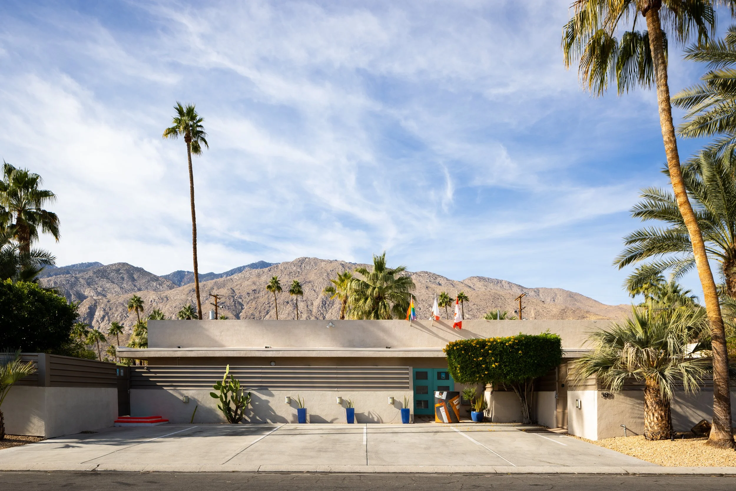 A desert landscape with palm trees in front of a building, mountains in the background, and a partly cloudy sky.