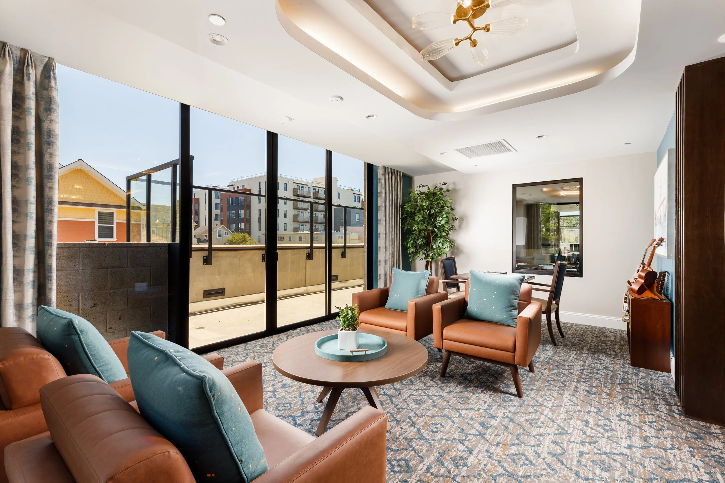 Bright living room with large sliding glass doors opening to a balcony, brown leather sofas with turquoise cushions, a round wooden coffee table with a small plant and decorative tray, patterned carpet, large potted plant, dining table with chairs, w