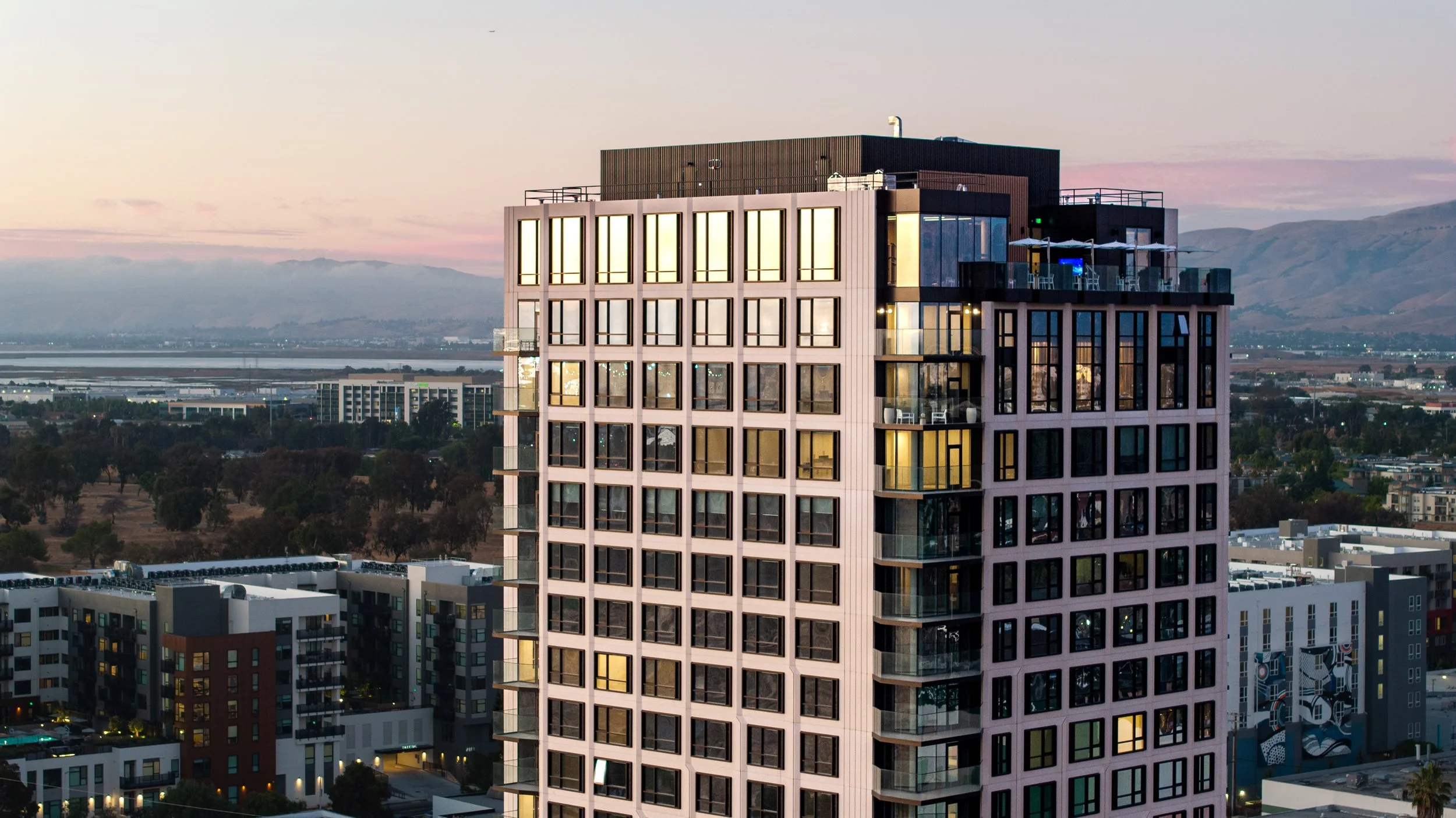 Modern high-rise building with illuminated windows against a sunset sky in an urban area.