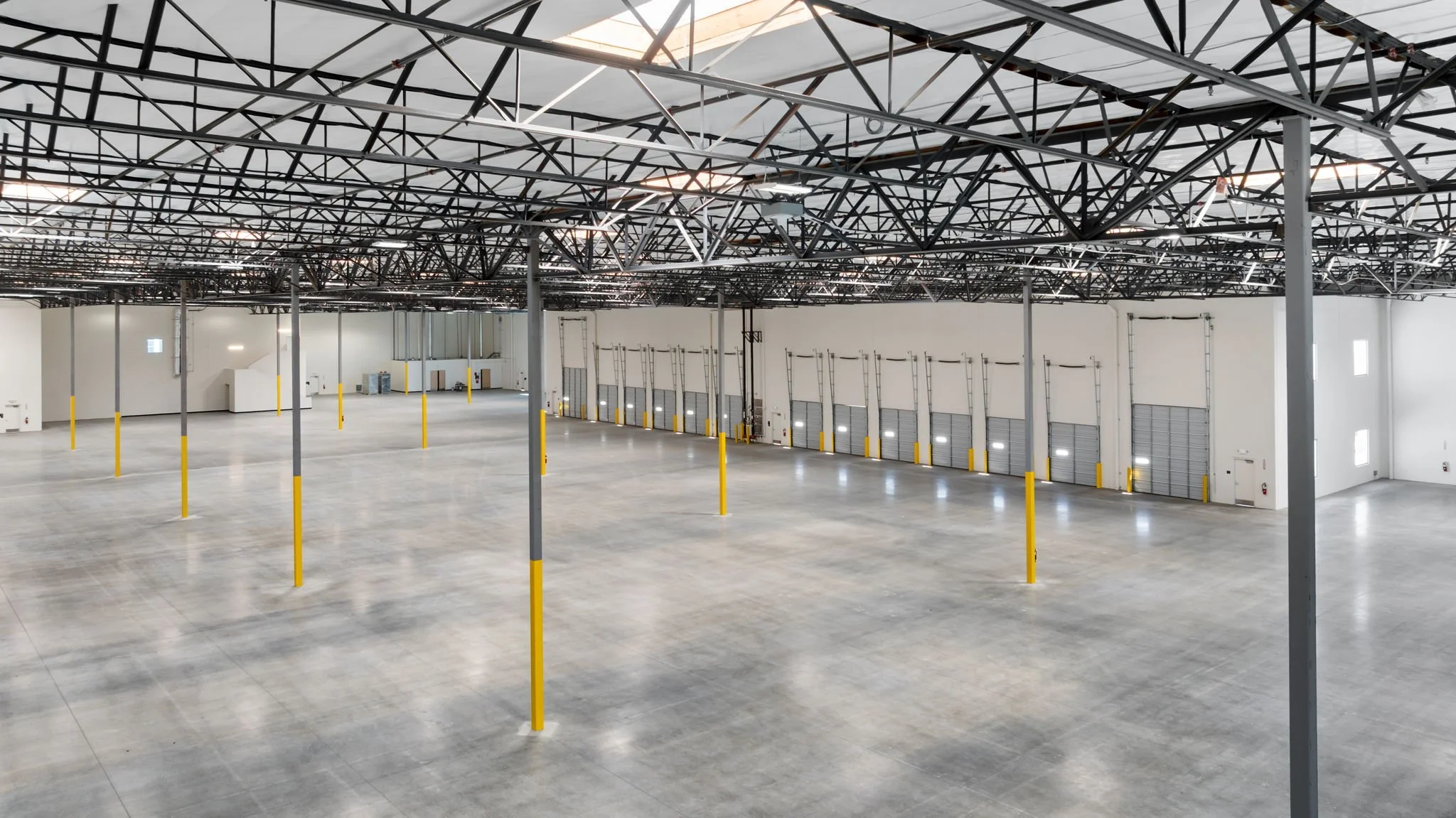 Empty warehouse interior with high ceilings, metal support beams, and yellow safety poles, with large garage doors along one wall.