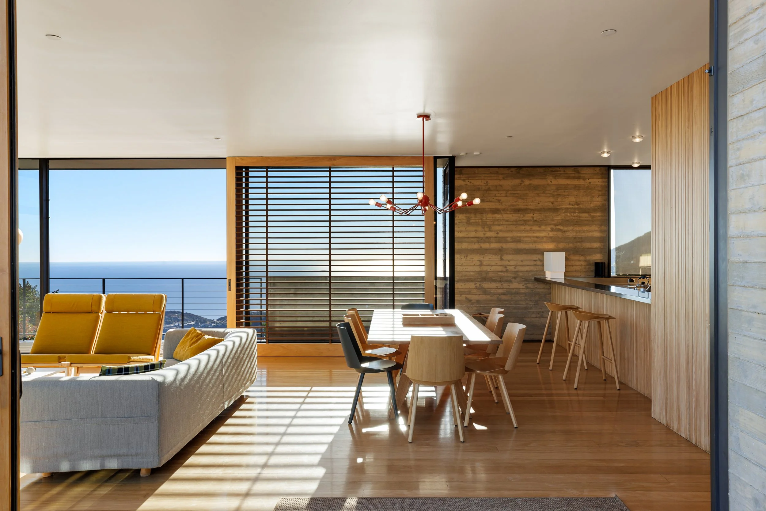 Interior of a modern living and dining area with wooden floors, a sofa with yellow cushions, a wooden dining table with mixed chairs, a light fixture, large windows revealing a view of the ocean, and a kitchen counter with barstools.