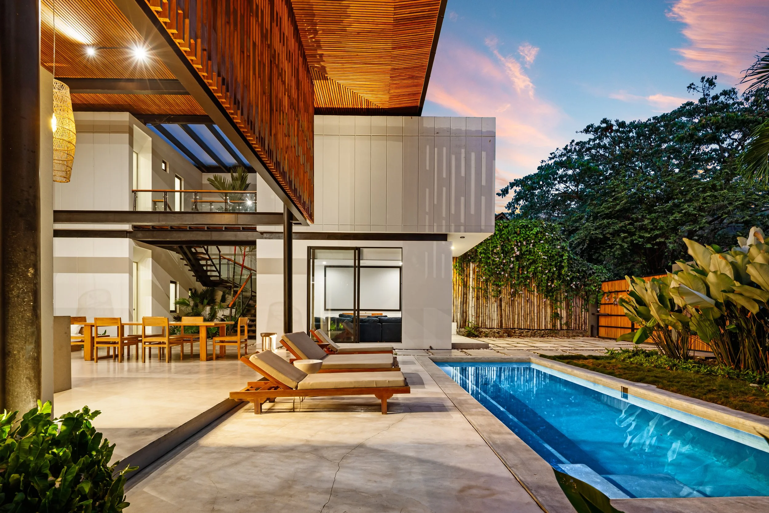 Modern house with a swimming pool, outdoor lounge chairs, and surrounding greenery at sunset.