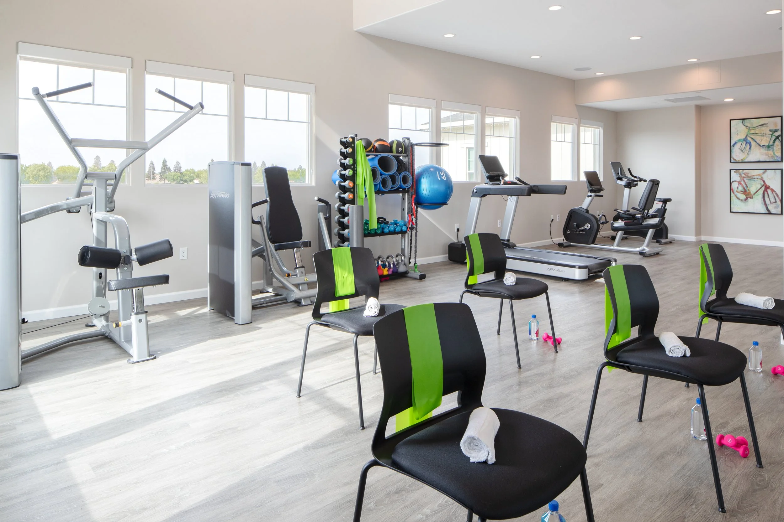 Bright, modern gym with cardio and strength equipment, chairs with towels, water bottles, and small weights, large windows, and colorful bike artwork on the walls.