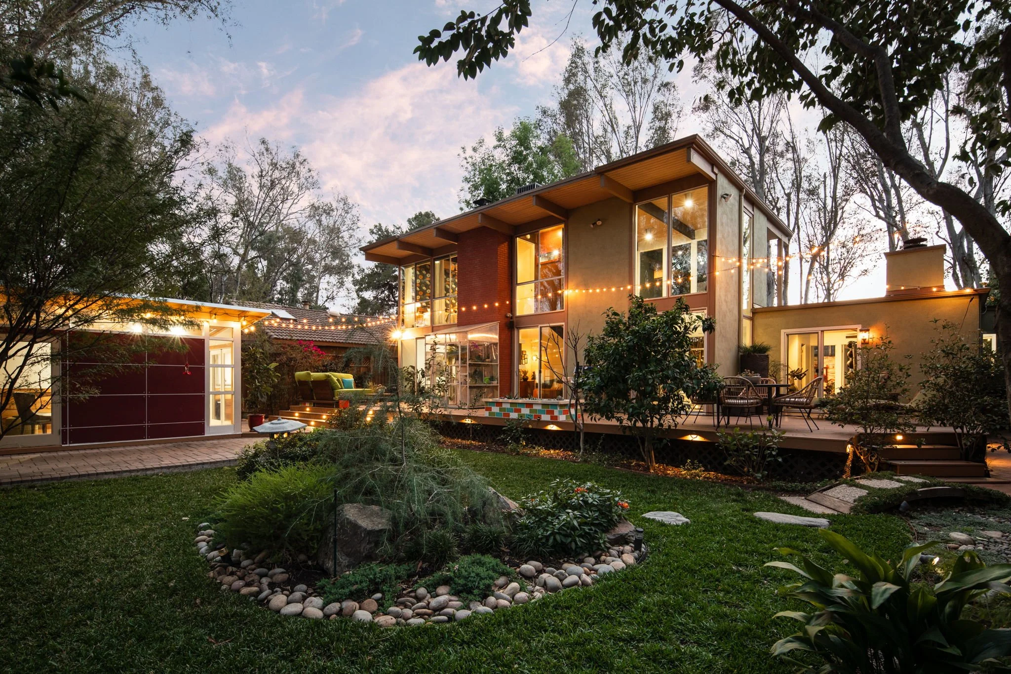 A modern two-story house with large windows, outdoor patio furniture, and string lights in a landscaped backyard at dusk.