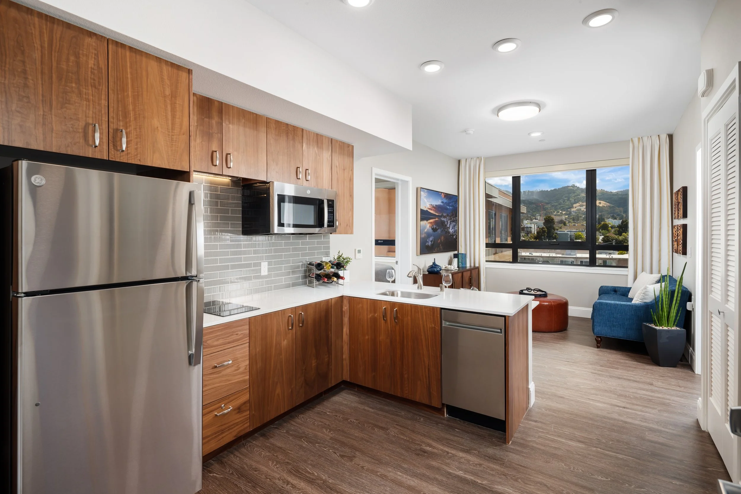 Modern kitchen with wooden cabinets, stainless steel refrigerator, microwave, and a white countertop with a sink, overlooking a living area with a blue sofa and a large window showing a cityscape and hills.