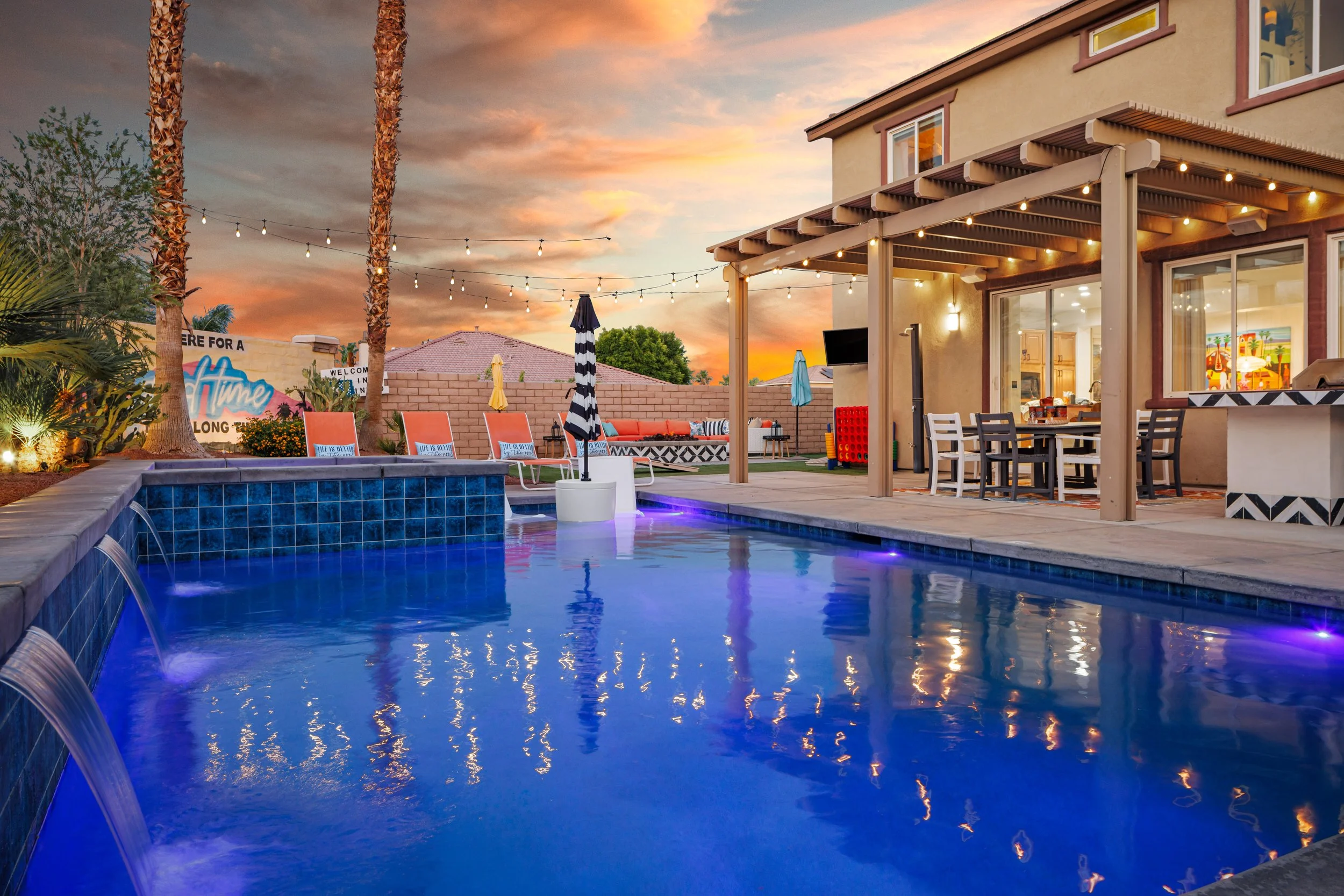 A backyard swimming pool illuminated at dusk with blue lighting, surrounded by poolside chairs with pillows, palm trees, string lights, a shaded patio area with a dining table and chairs, and an outdoor seating area with a sofa and umbrellas, with a sunset sky in the background.