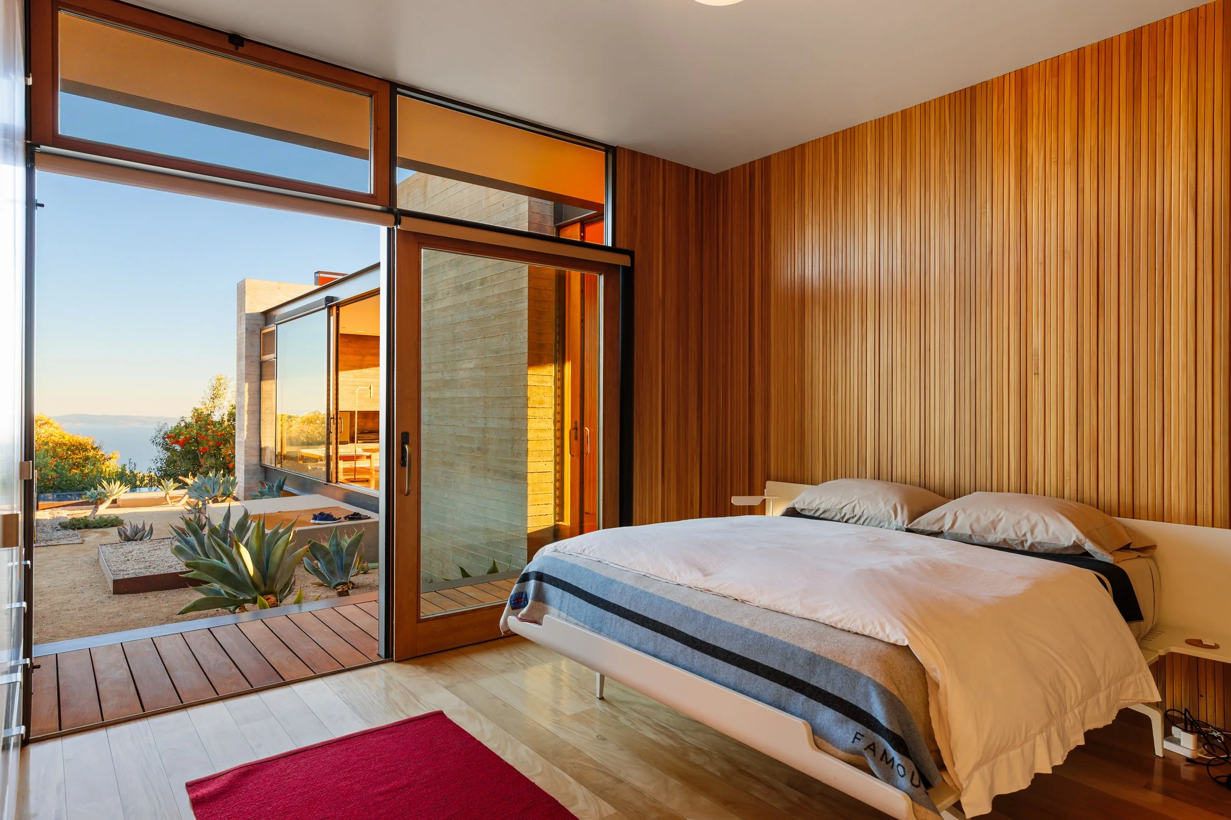 A modern bedroom with a wooden-paneled accent wall, a large bed with white sheets and pillows, and a sliding glass door leading to a balcony with plants and a scenic view of trees and water.