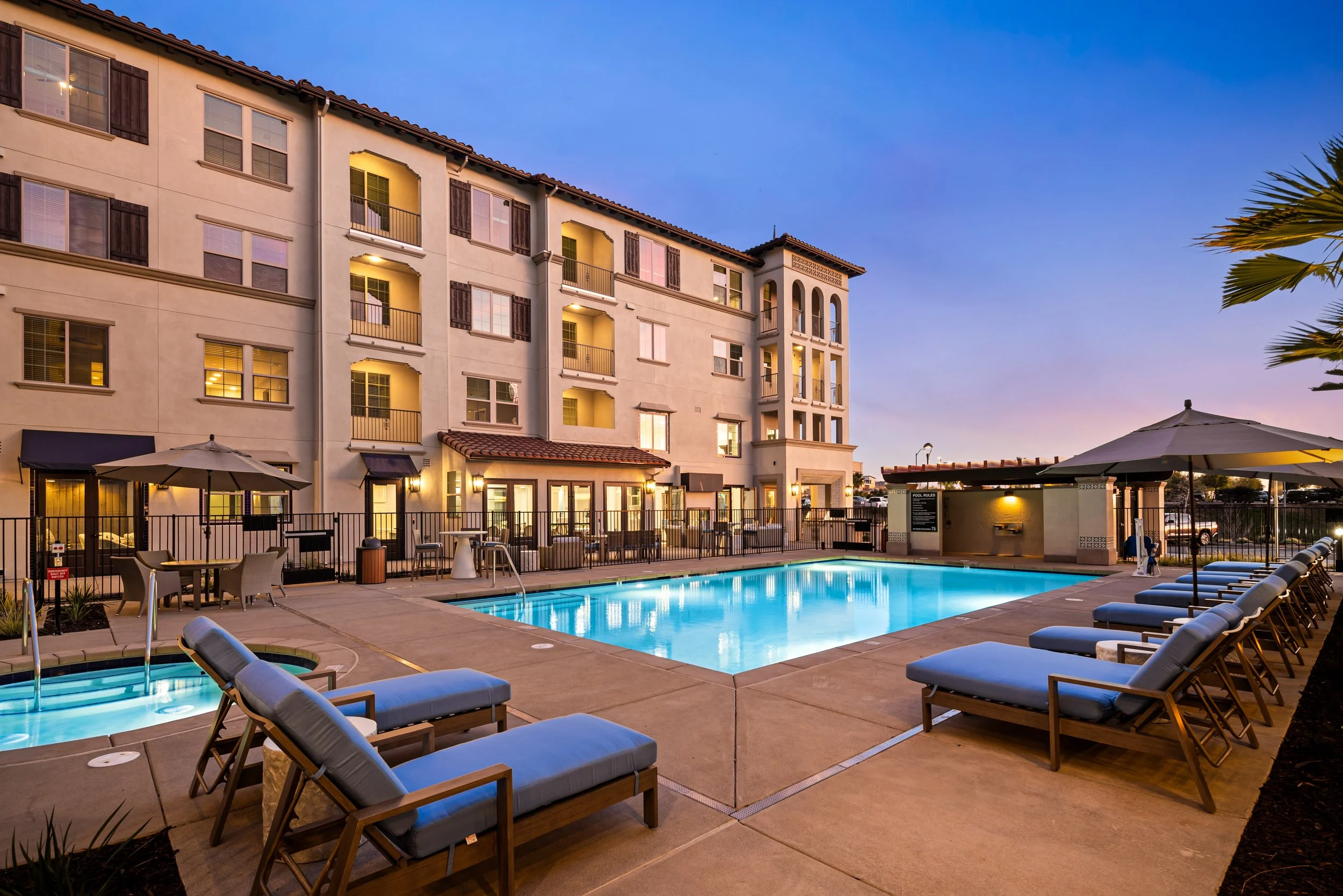 An outdoor swimming pool area at sunset, surrounded by lounge chairs, umbrellas, and a multi-story residential or hotel building with balconies and lit windows.