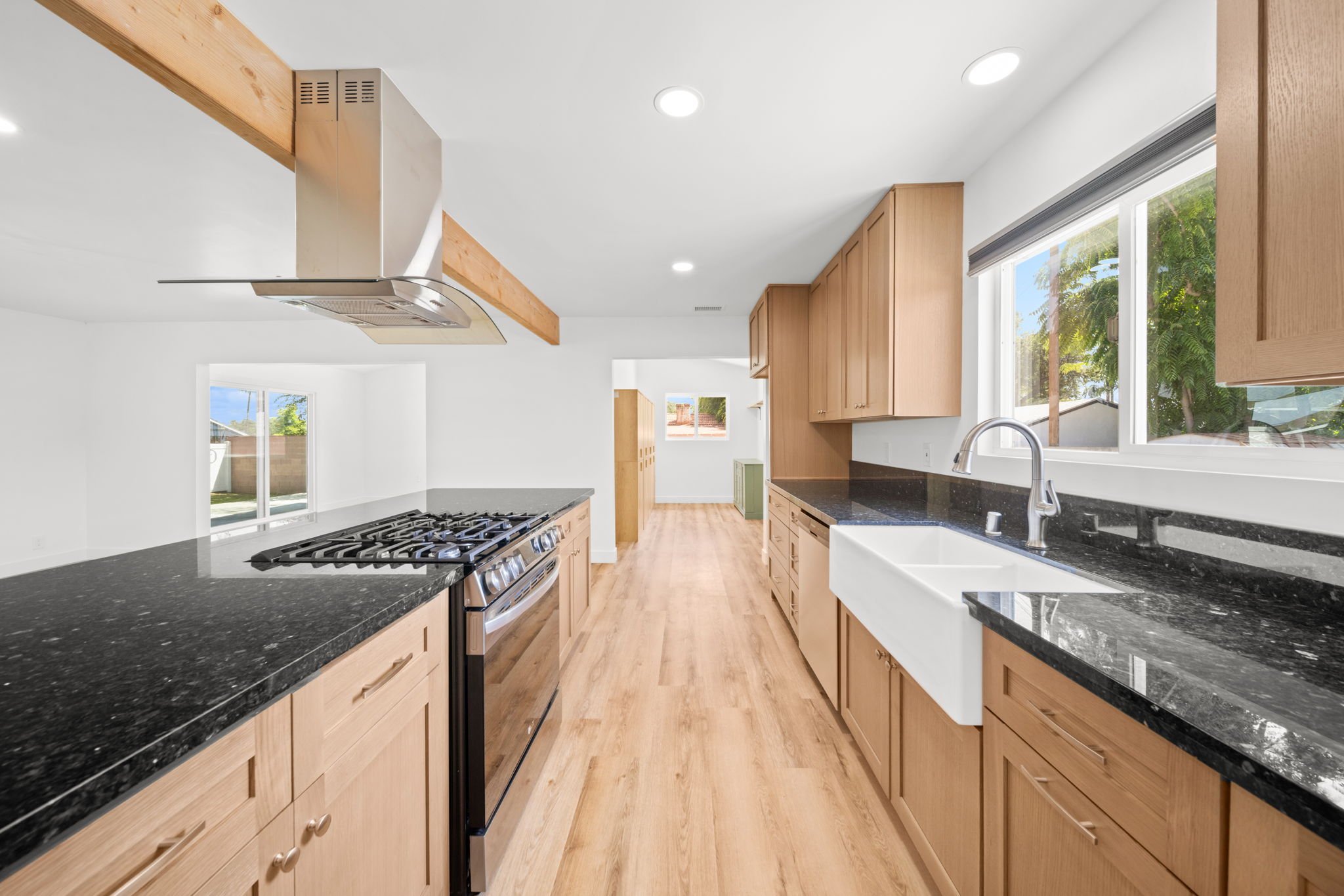 Modern kitchen with wooden cabinets, black granite countertops, stainless steel stove and range hood, large window with a view of trees, bright white walls and ceiling with recessed lighting, light wood flooring.