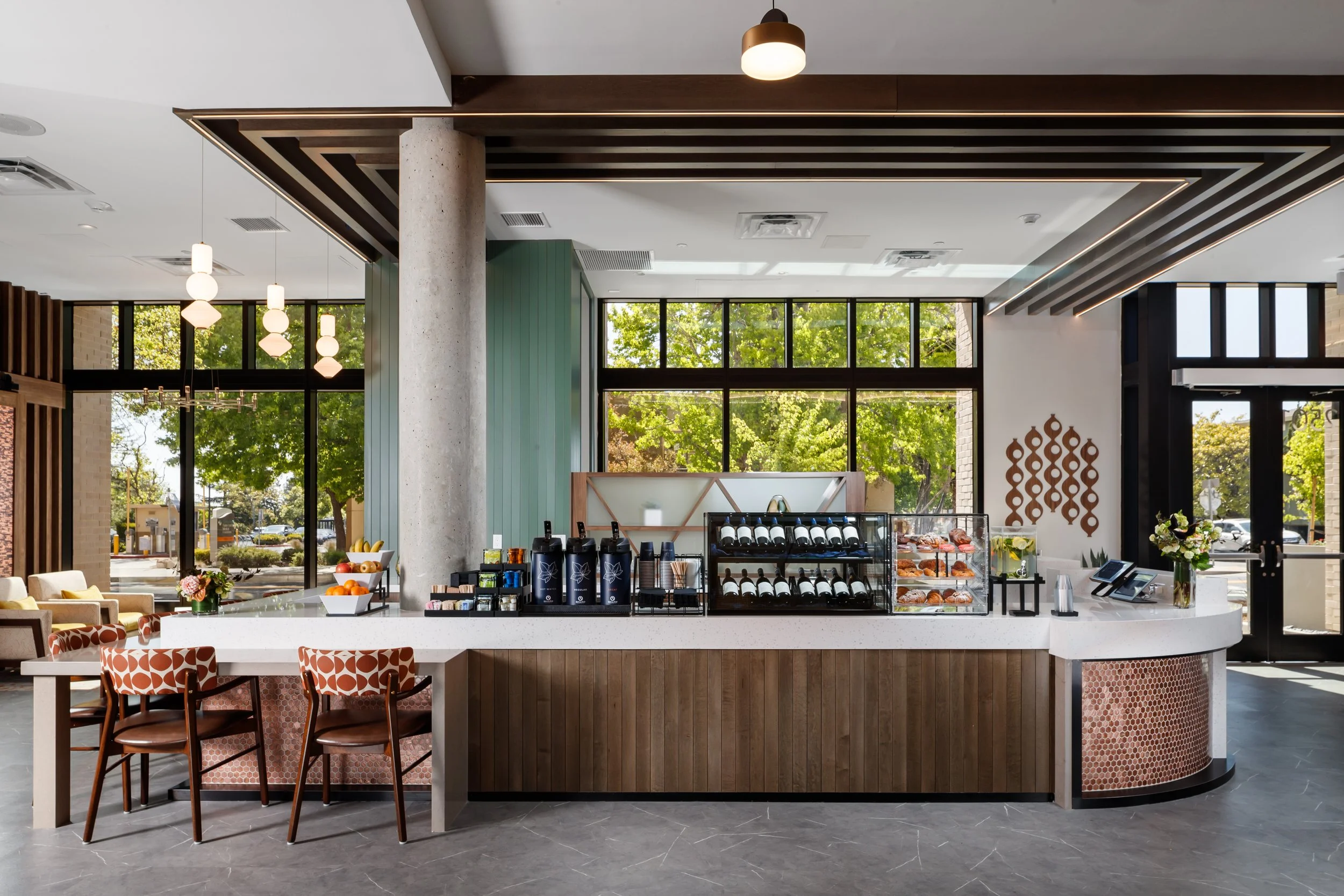 Modern café counter with beverages, pastries, and fruit, in a bright space with large windows showing green trees outside.