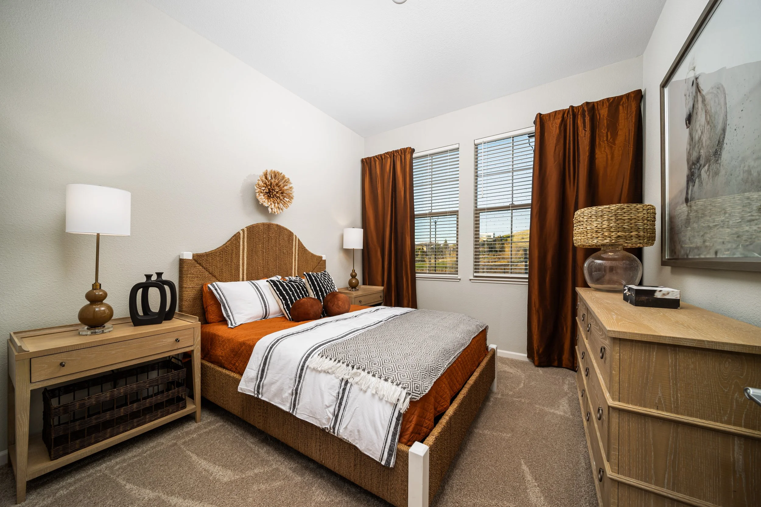 A bedroom with a bed, two nightstands, and a dresser with a lamp, artwork, and window blinds.