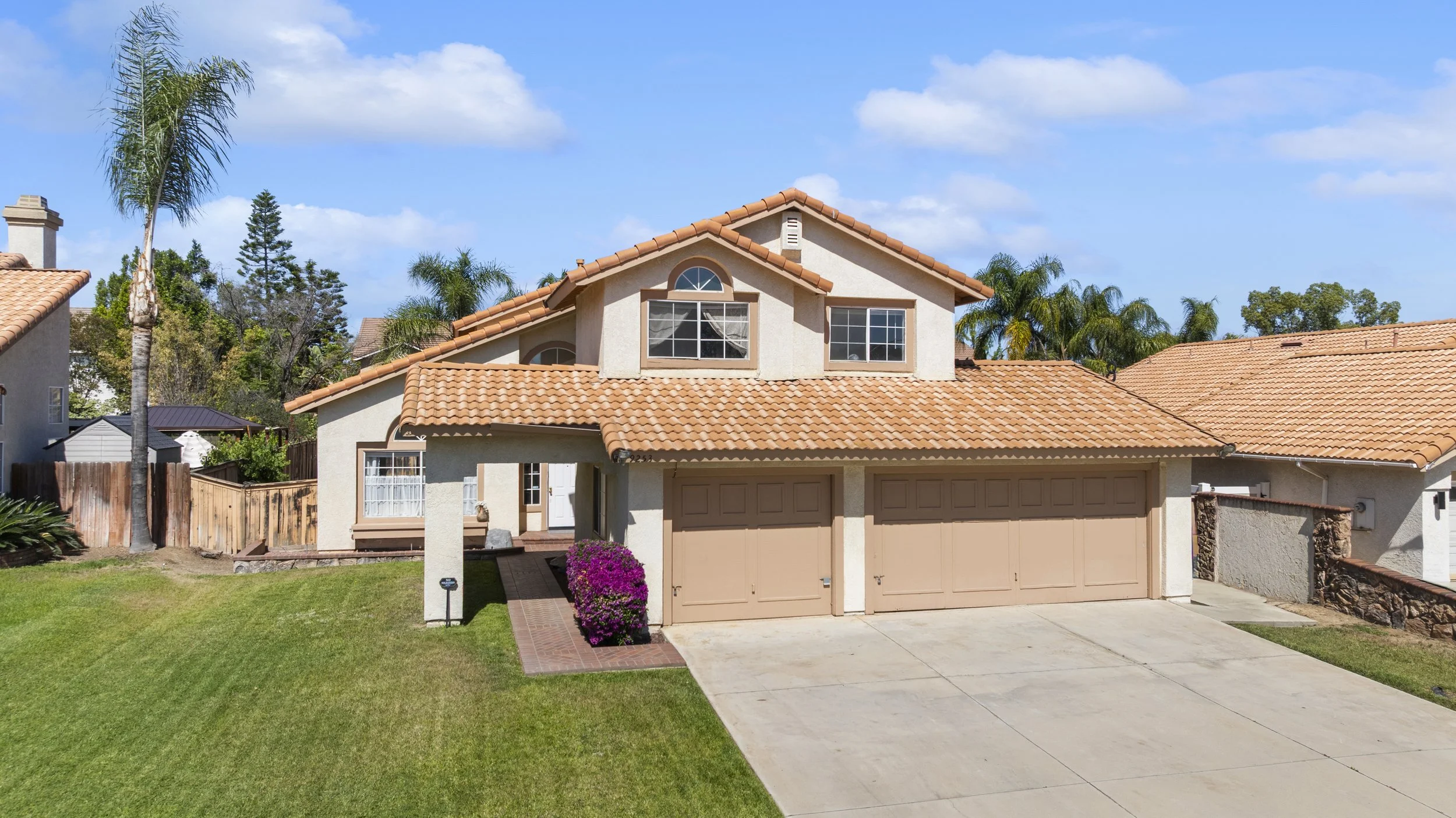Two-story suburban house with a terracotta tiled roof, beige garage doors, and a front lawn with green grass and pink flowers. Palm trees and other trees are visible in the background under a partly cloudy sky.