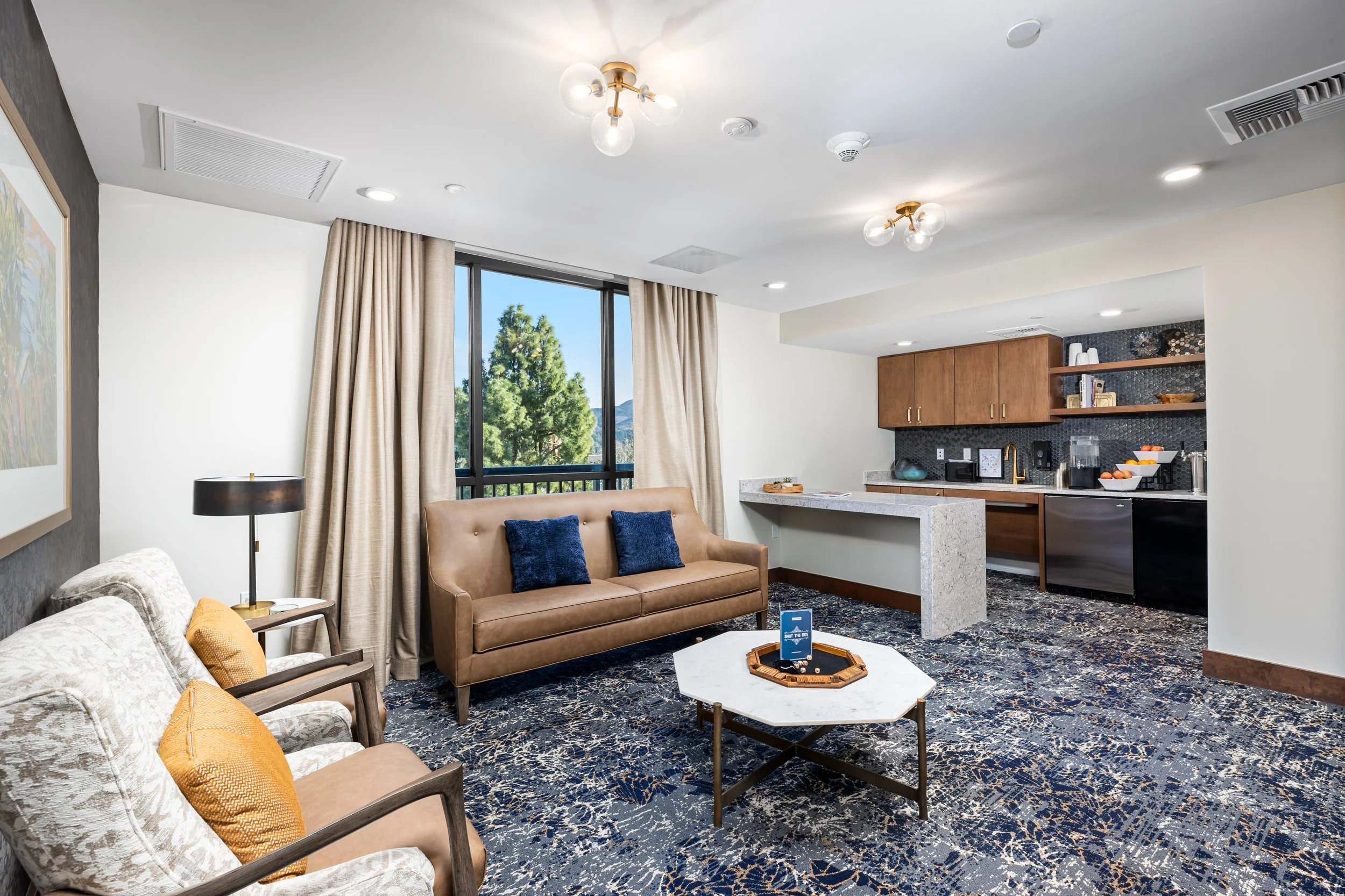 Living room with beige and white armchairs, a brown sofa with blue pillows, a marble coffee table, a large window with beige curtains, a lighting fixture on the ceiling, a kitchenette with wooden cabinets, black backsplash, and open shelves with kitc