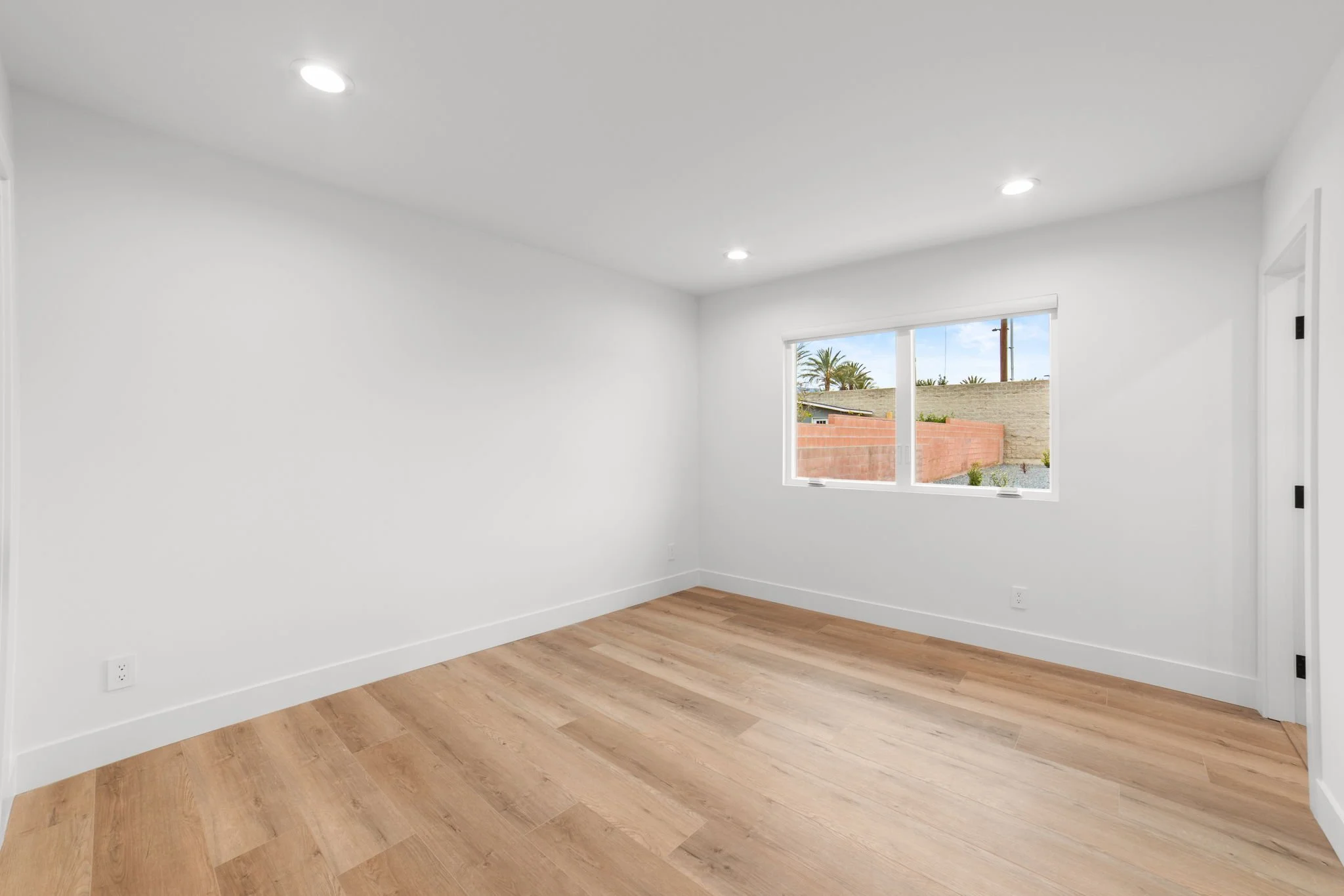 Empty room with white walls, hardwood flooring, a large window showing a brick wall and palm trees outside, and recessed ceiling lights.