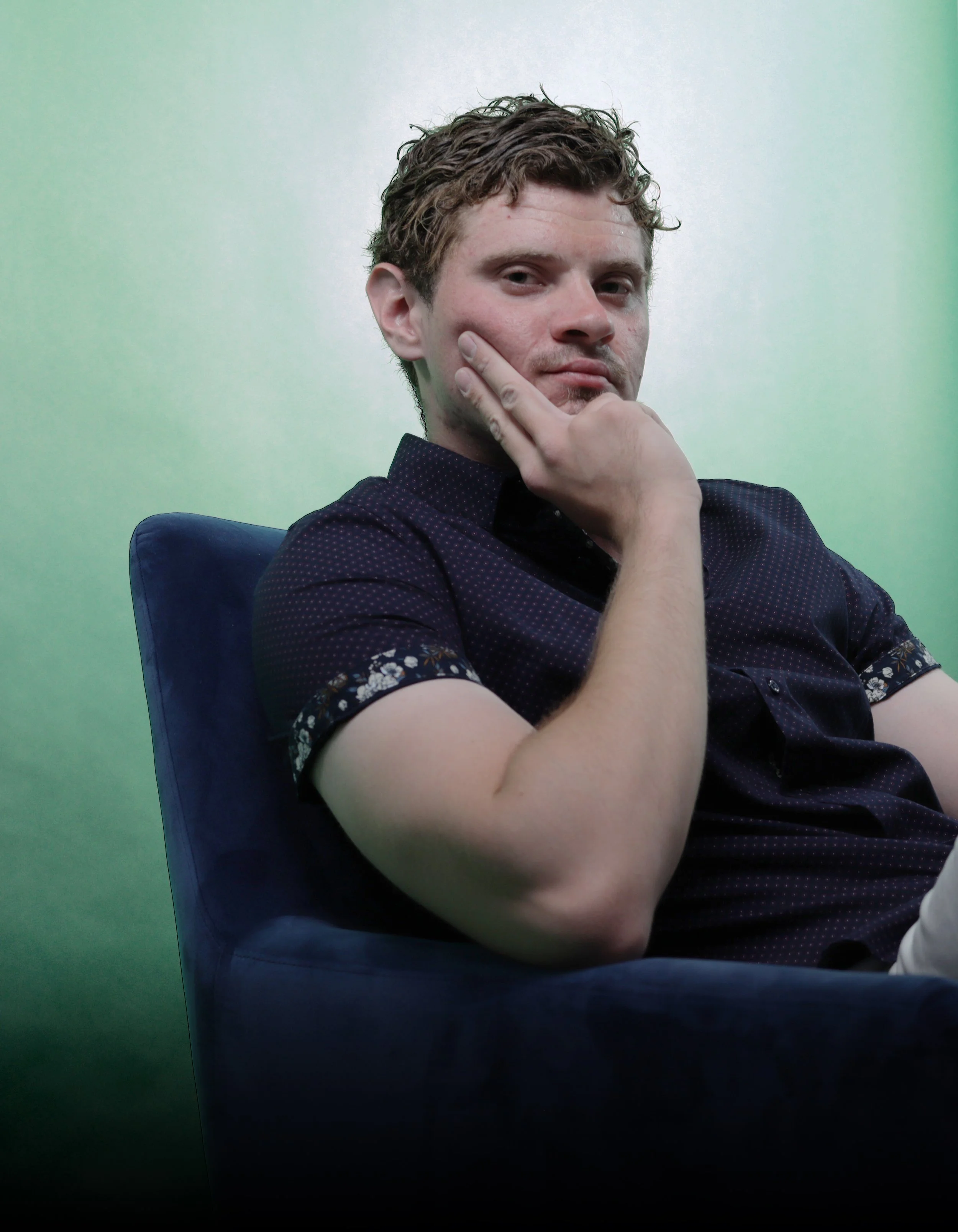 A young man with curly brown hair, wearing a dark shirt with a floral pattern on the sleeves, sitting in a blue chair against a green background, making a thoughtful pose with his hand on his chin and fingers on his face.