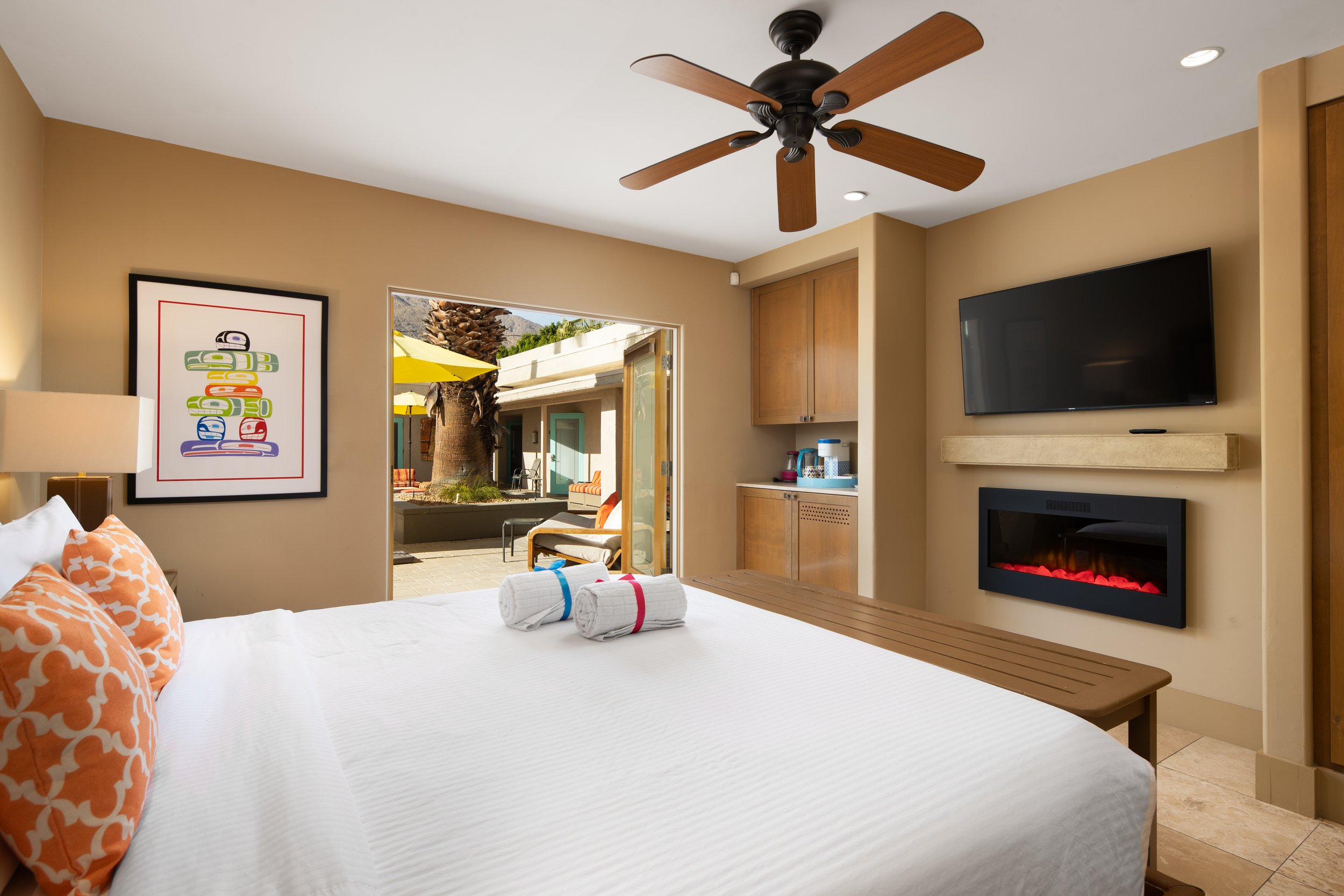Hotel room with a king-size bed, orange patterned pillows, towels on the bed, a framed colorful art piece on the wall, a wall-mounted television, an electric fireplace, wooden furniture, ceiling fan, and an open sliding door leading to an outdoor pat