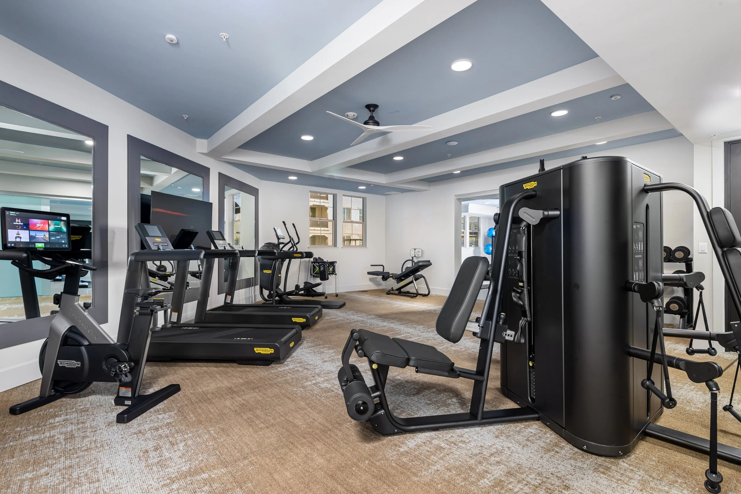 Indoor gym with treadmills, stationary bikes, and weight equipment in a well-lit room with large windows and a ceiling fan.