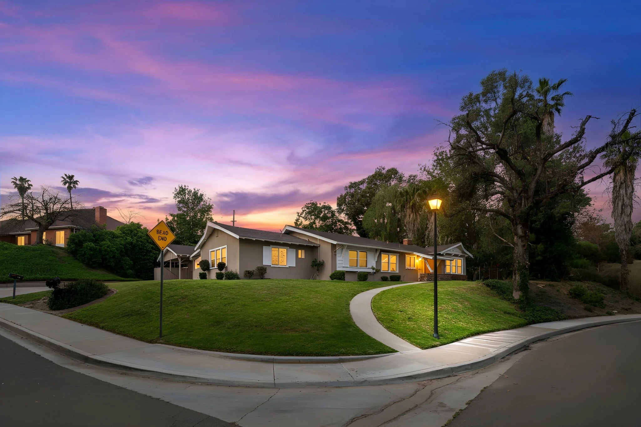 A view of a neighborhood at dusk with a purple and pink sky, a house with illuminated windows, a streetlamp, and green lawns.