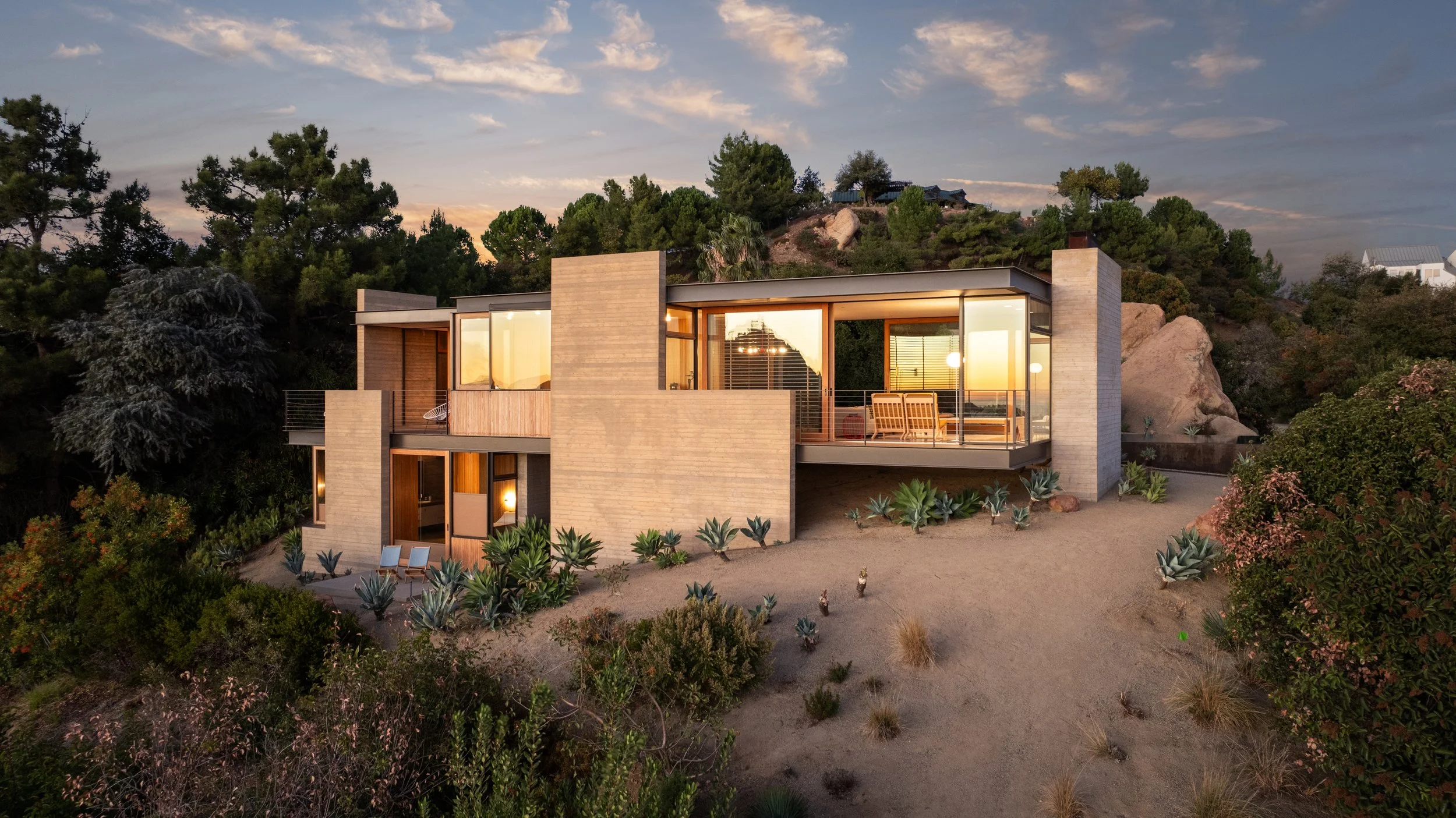 Modern house on hillside with large windows, surrounded by desert plants and trees, during sunset.
