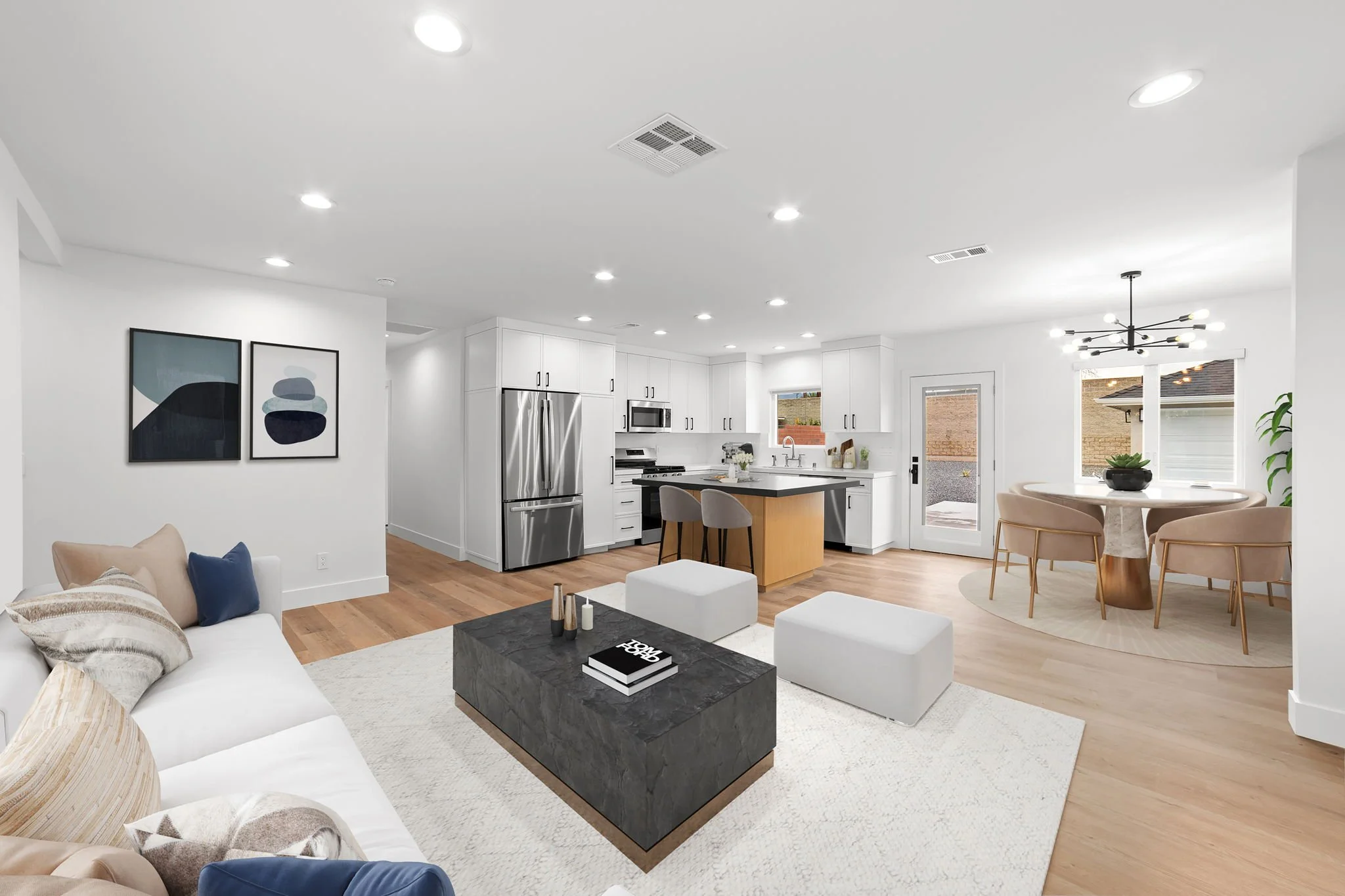 A modern open-concept living and kitchen area with white walls and wood flooring. The space includes a white sofa with pillows, a black marble coffee table, and artwork on the wall. The kitchen features stainless steel appliances, white cabinets, and an island with bar stools. There is a round dining table with chairs, a geometric light fixture, and large windows bringing in natural light.