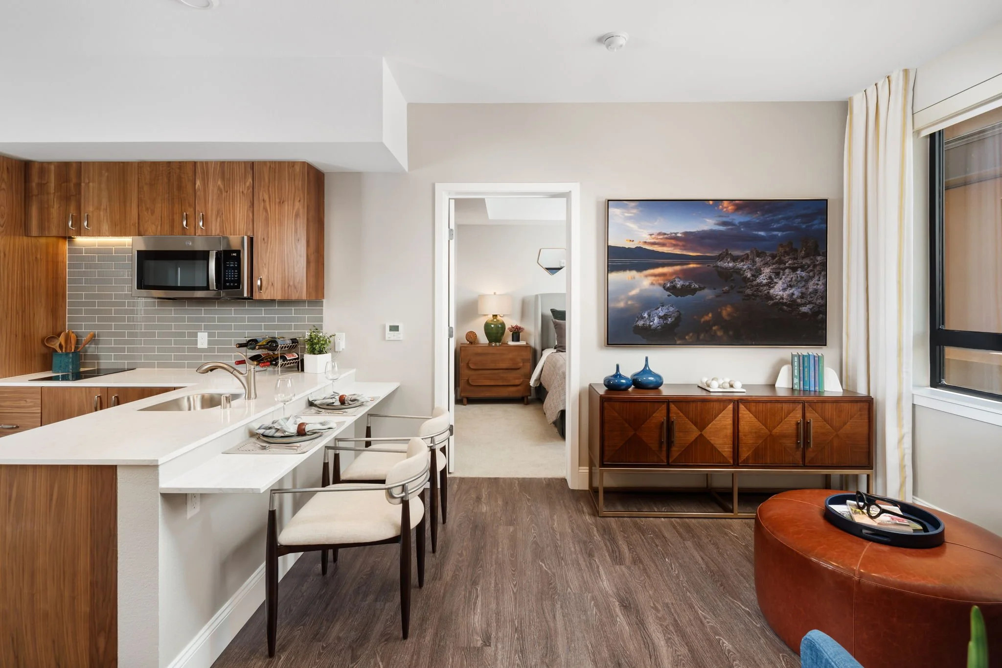 Interior of a modern apartment with a kitchen and living room. The kitchen features wooden cabinets, a microwave, and a countertop with dining plates and glasses. The living room has a large wall-mounted TV displaying a nature scene, a wooden sideboard with blue vases, and a window with cream curtains. There is a glimpse of a bedroom with a bed, nightstand, and lamp through an open doorway.