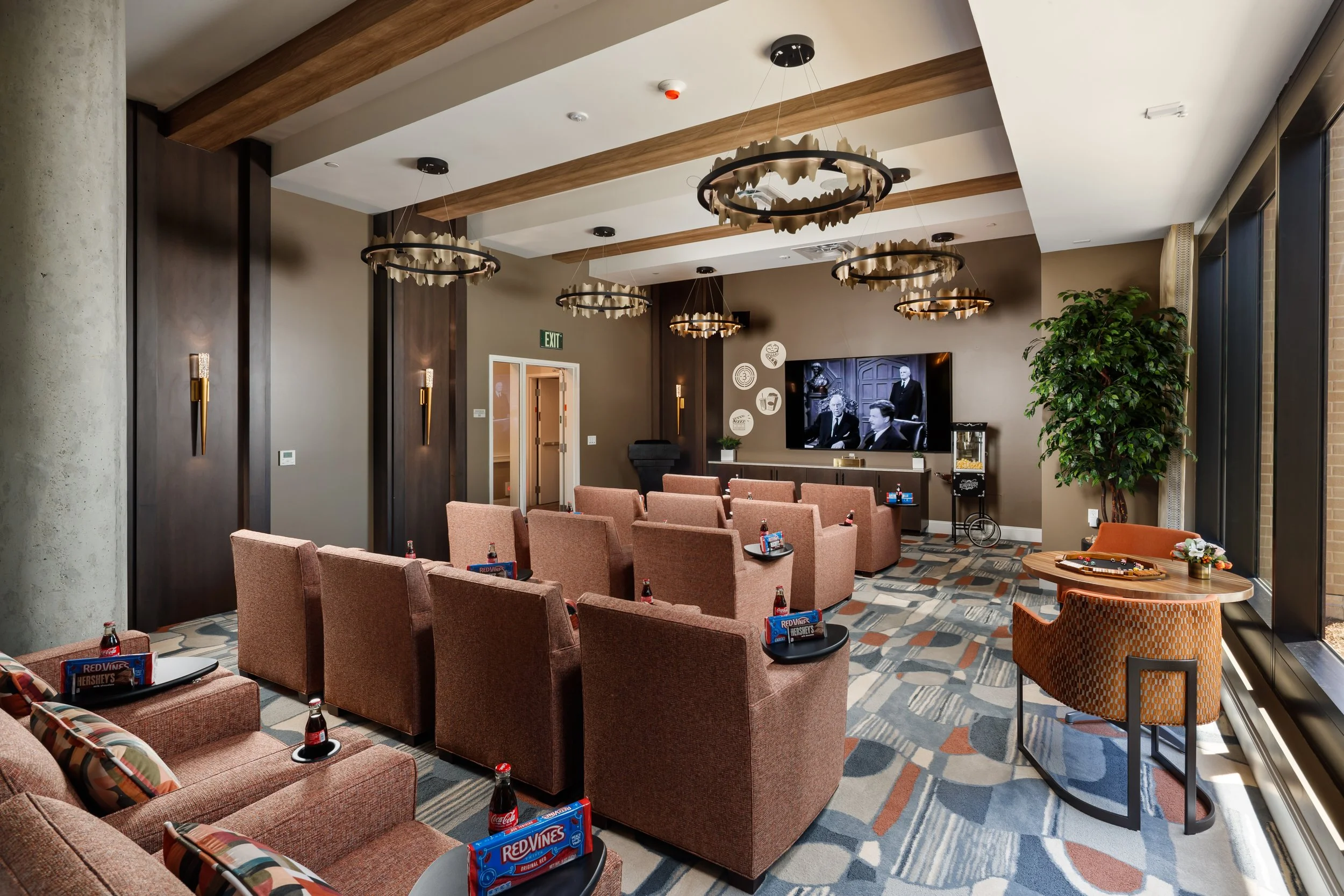 A modern lounge area with pink upholstered chairs arranged in rows, each with a tray of snacks and drinks. There is a large flat-screen TV on the wall, a popcorn machine, a round table with chairs, and a large potted plant near the window. The room h