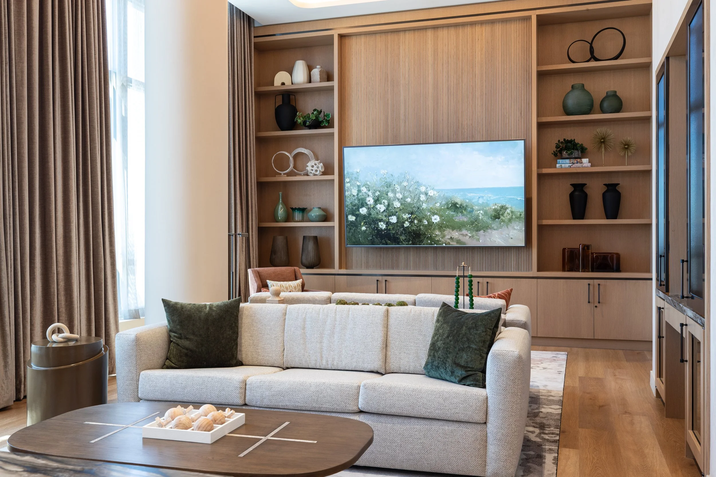 Modern living room with a large wall-mounted TV, beige sofa with dark green pillows, round wooden coffee table with a white tray, built-in wooden shelving with decorative vases and art, and brown curtains.