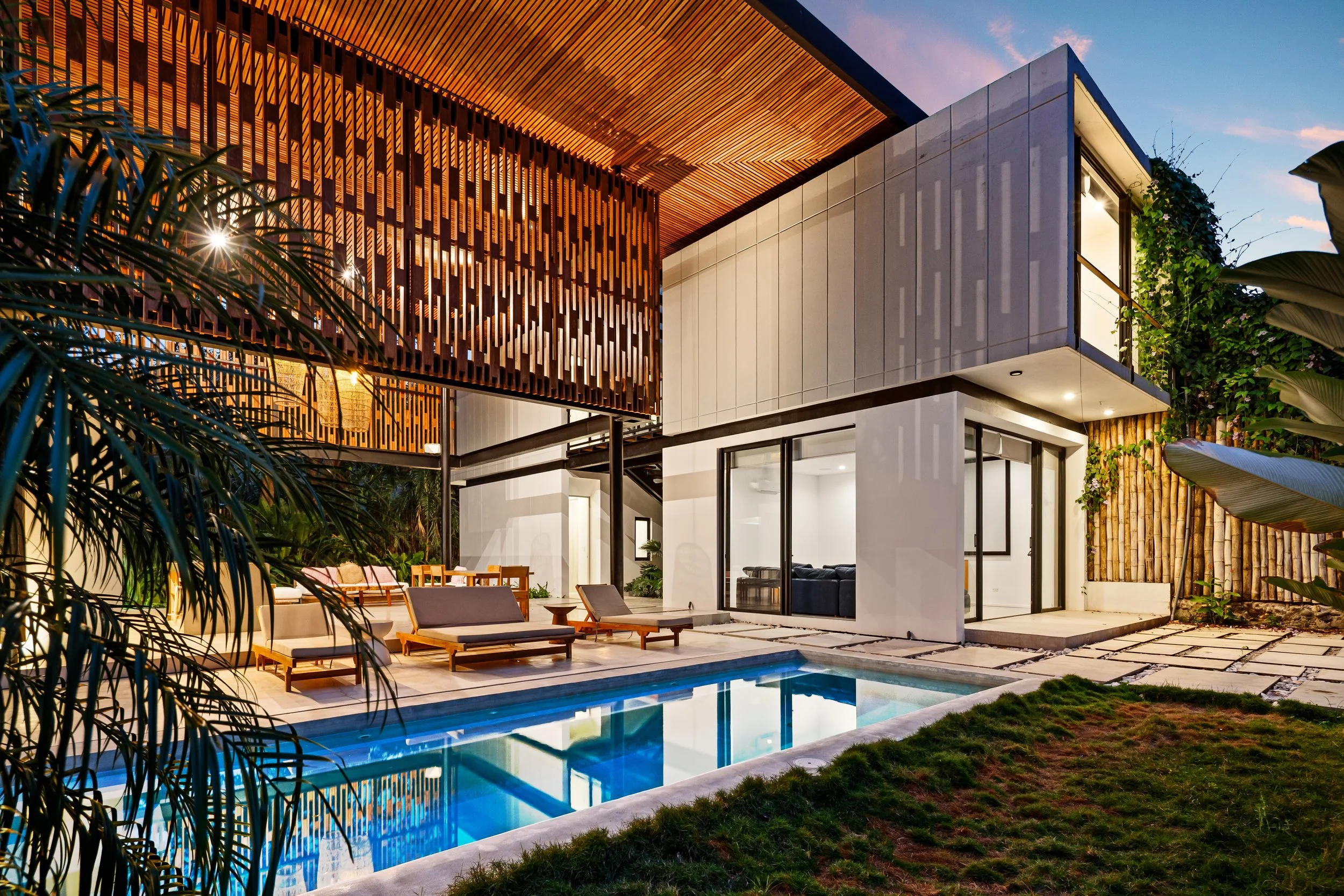 Modern two-story house with outdoor swimming pool, patio furniture, and lush greenery at dusk