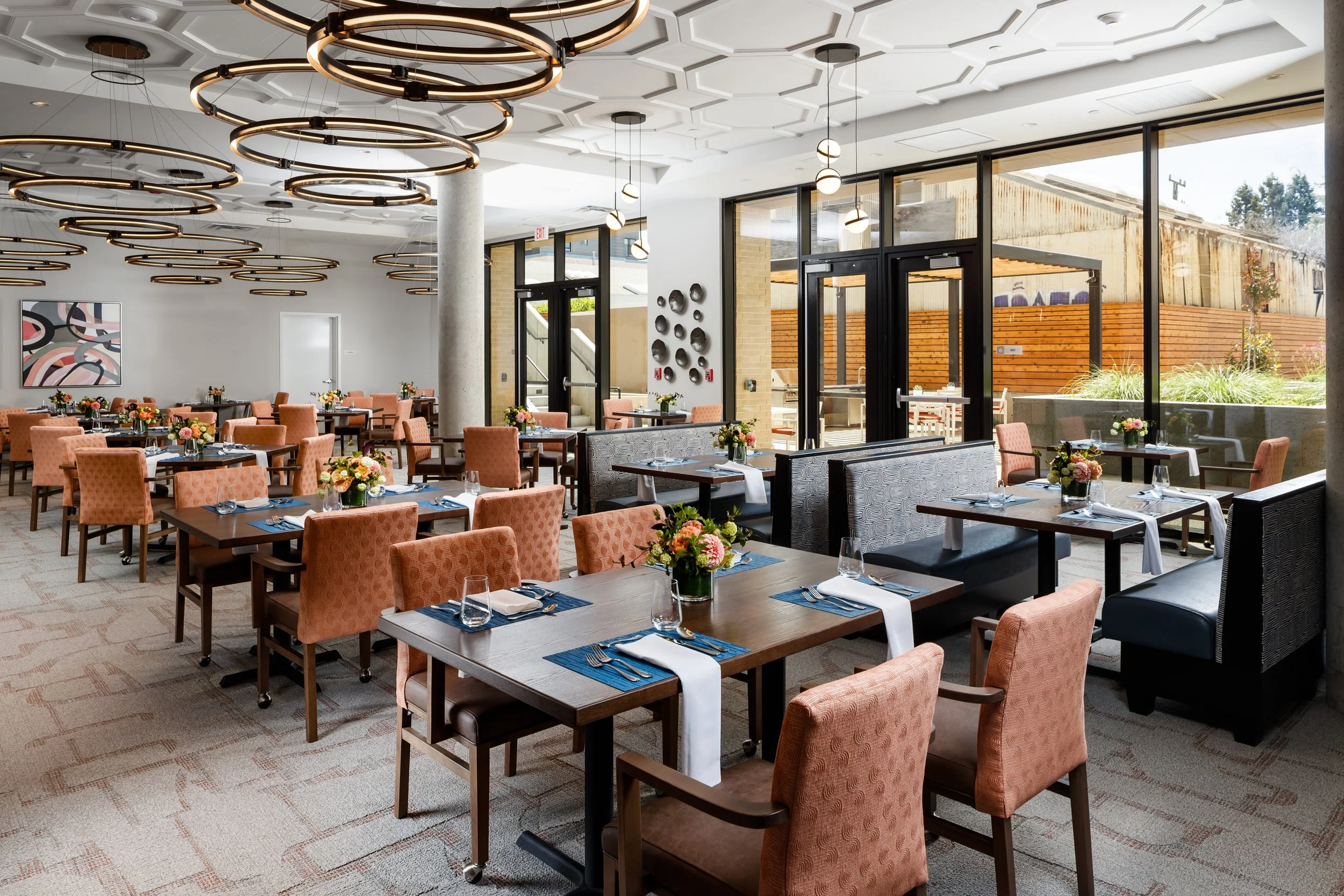 A spacious, brightly lit restaurant with modern decor. Tables are set with blue placemats, white napkins, glasses, and floral centerpieces. Peach-colored chairs and grey banquettes surround the tables. Large glass windows and doors reveal an outdoor 