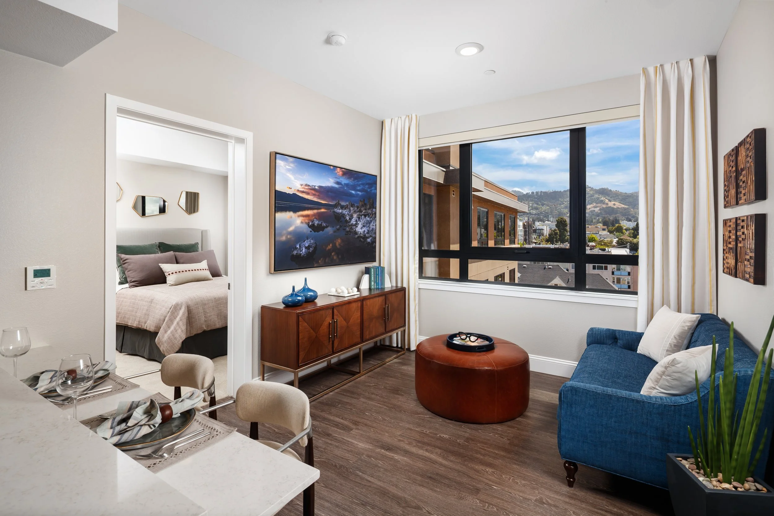 Living room with large window, blue sofa, brown ottoman, TV, dining table with chairs, and a view of city buildings and hills.