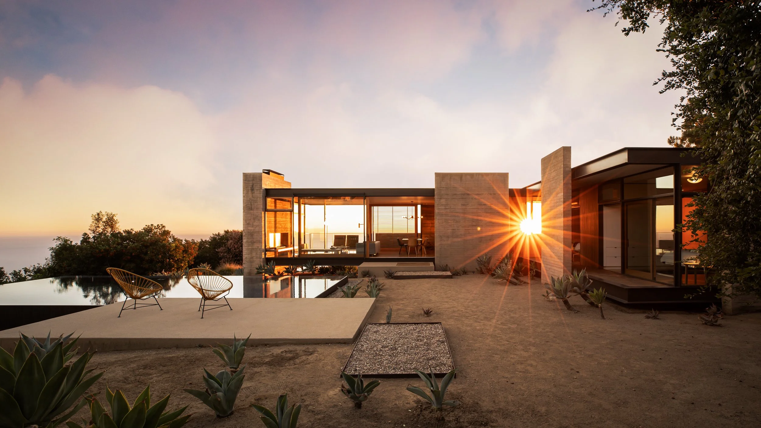 Modern house with large glass windows and outdoor seating, sunset sun rays visible, desert landscaping with succulents, hillside view, and clear sky.