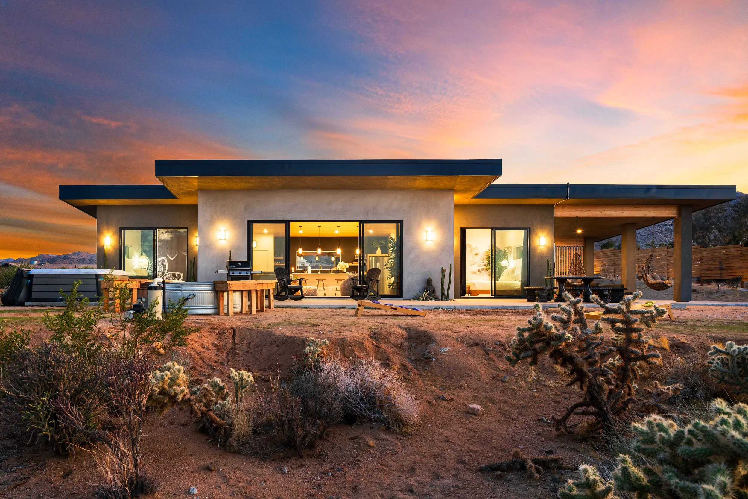 Modern one-story house in a desert landscape at sunset, with large glass sliding doors, an outdoor seating area, and cacti in the foreground.