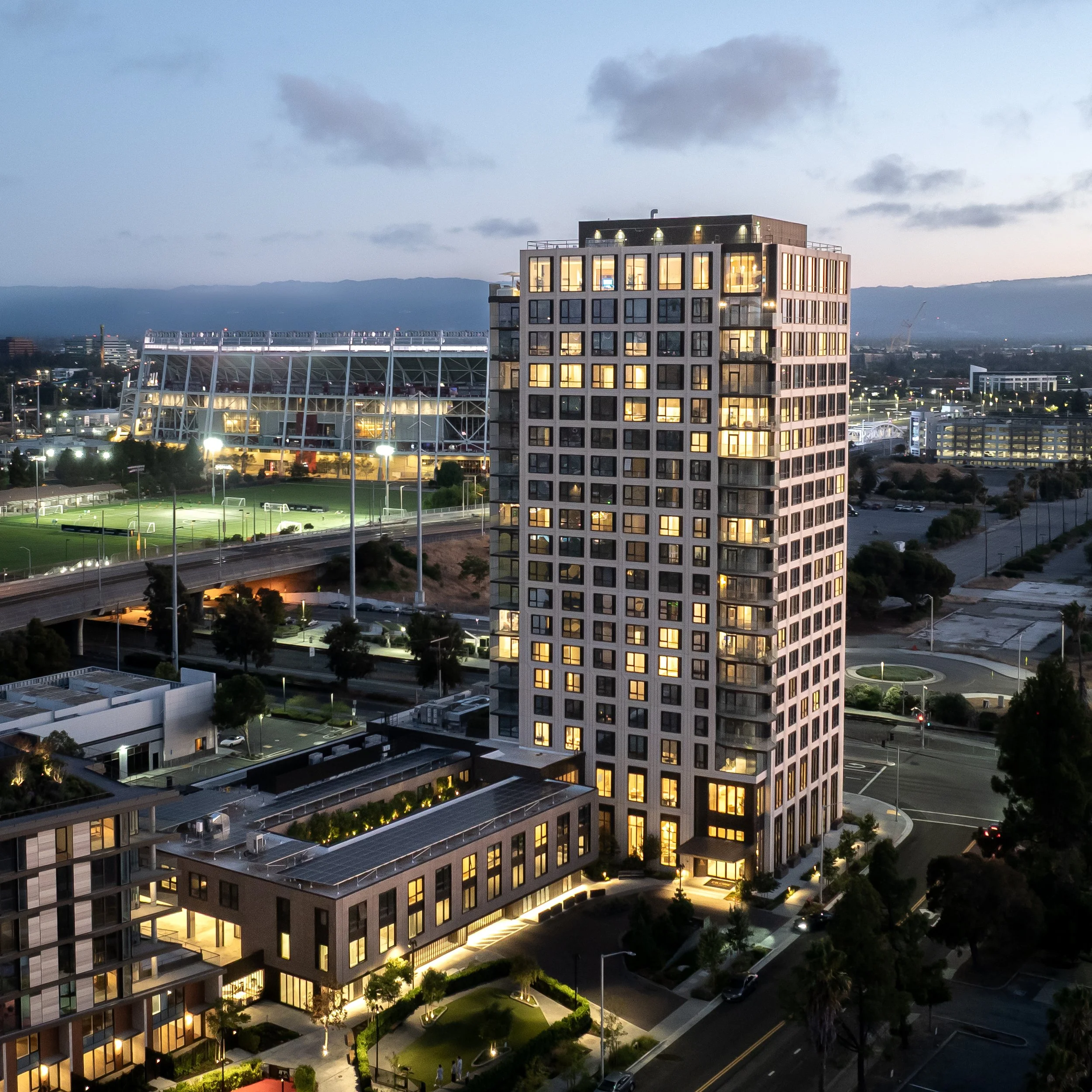 High-rise apartment building illuminated at dusk with cityscape and sports stadium in the background.