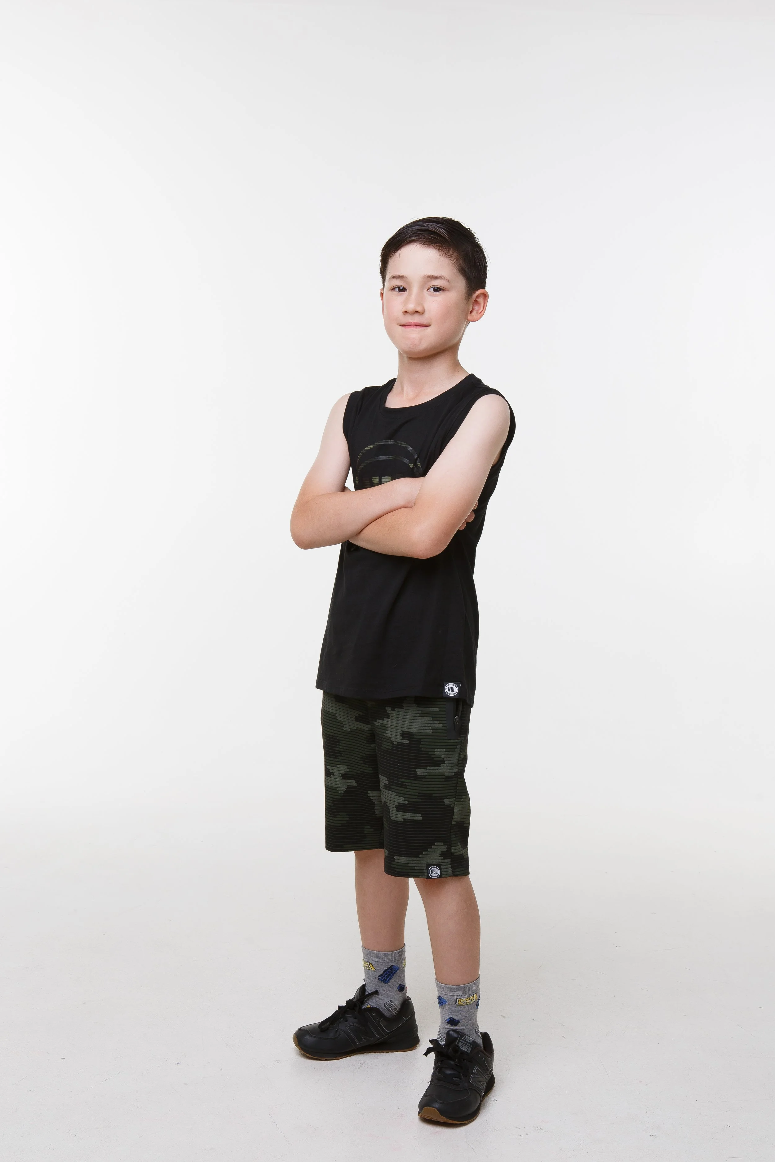 A young boy standing with arms crossed, wearing a black sleeveless shirt, camo shorts, gray socks with patterns, and black sneakers, against a plain white background.