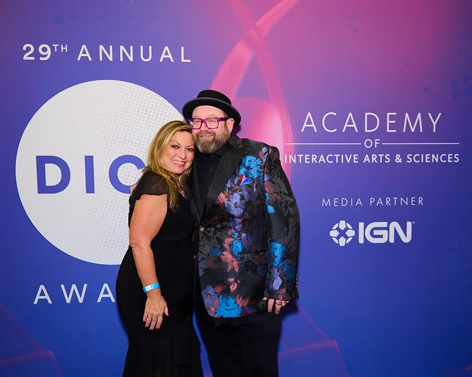 A man and woman posing together at the 29th Annual DICE Awards, in front of a blue and purple backdrop with event and media partner logos, smiling at the camera.