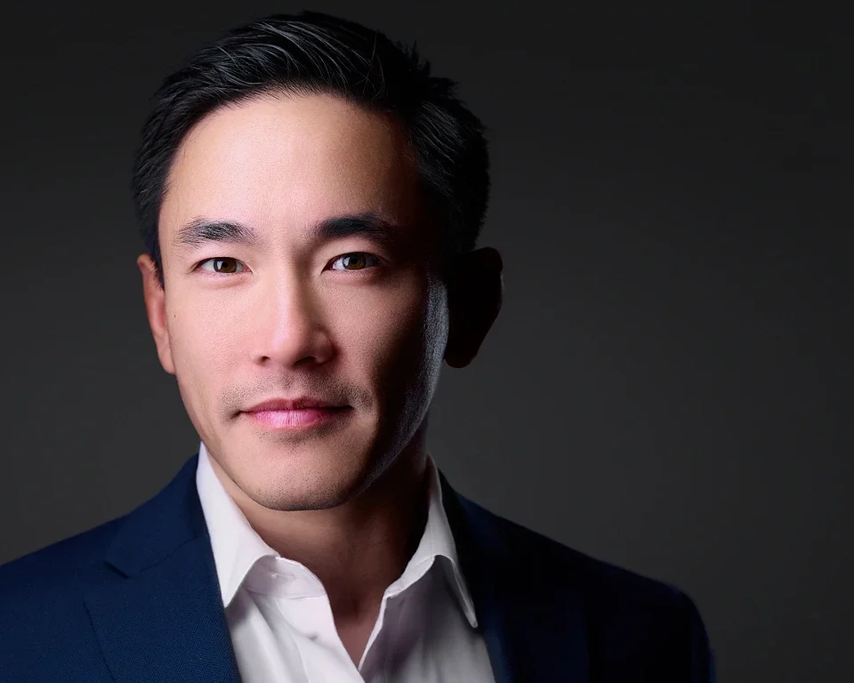 Portrait of a young Asian man in a dark suit and white shirt against a dark background.