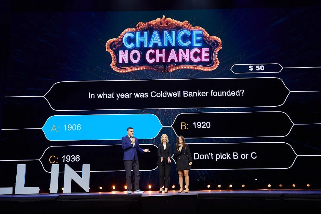 A game show stage with three hosts standing in front of a large screen displaying a question about the founding year of Coldwell Banker. The screen offers multiple choice answers: 1906, 1920, and 1936, with the highlighted answer being 1906. The show