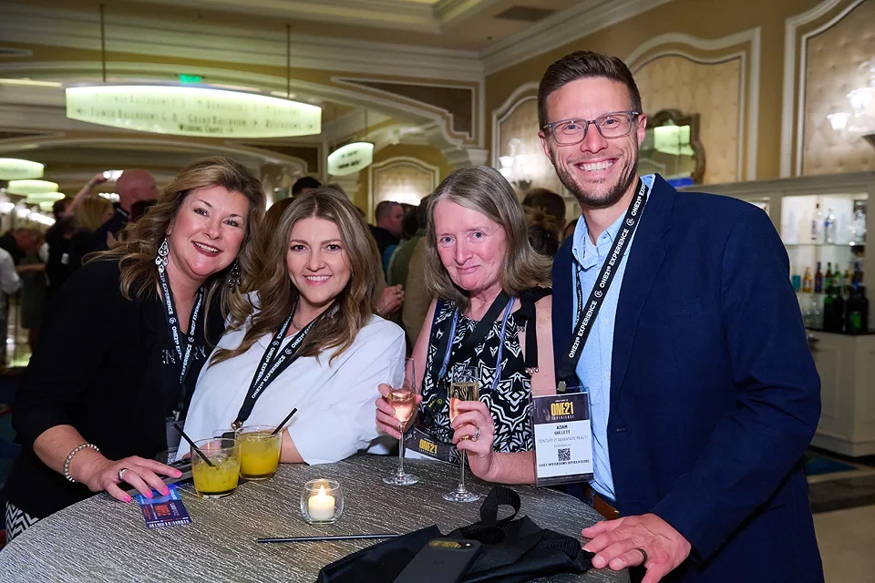 Four people smiling and holding drinks at a social event in a banquet hall, with a bar in the background.