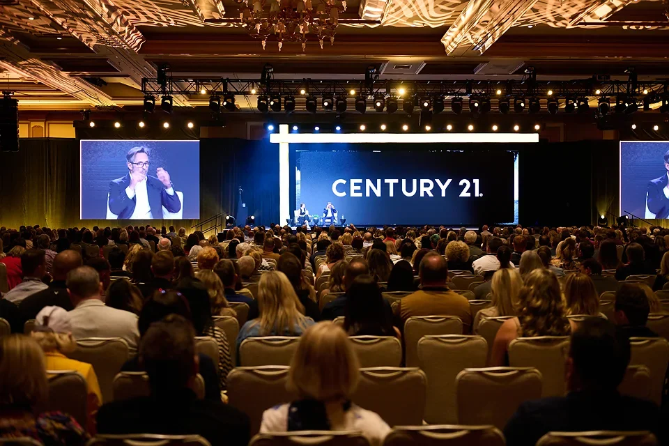 Large conference hall filled with seated audience watching a presentation on stage with a large screen displaying 'CENTURY 21'. A speaker is visible on the stage, and two large screens show the speaker.