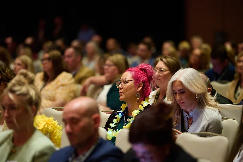 Audience at a conference listening attentively, with a woman with bright pink hair and glasses sitting in the middle.