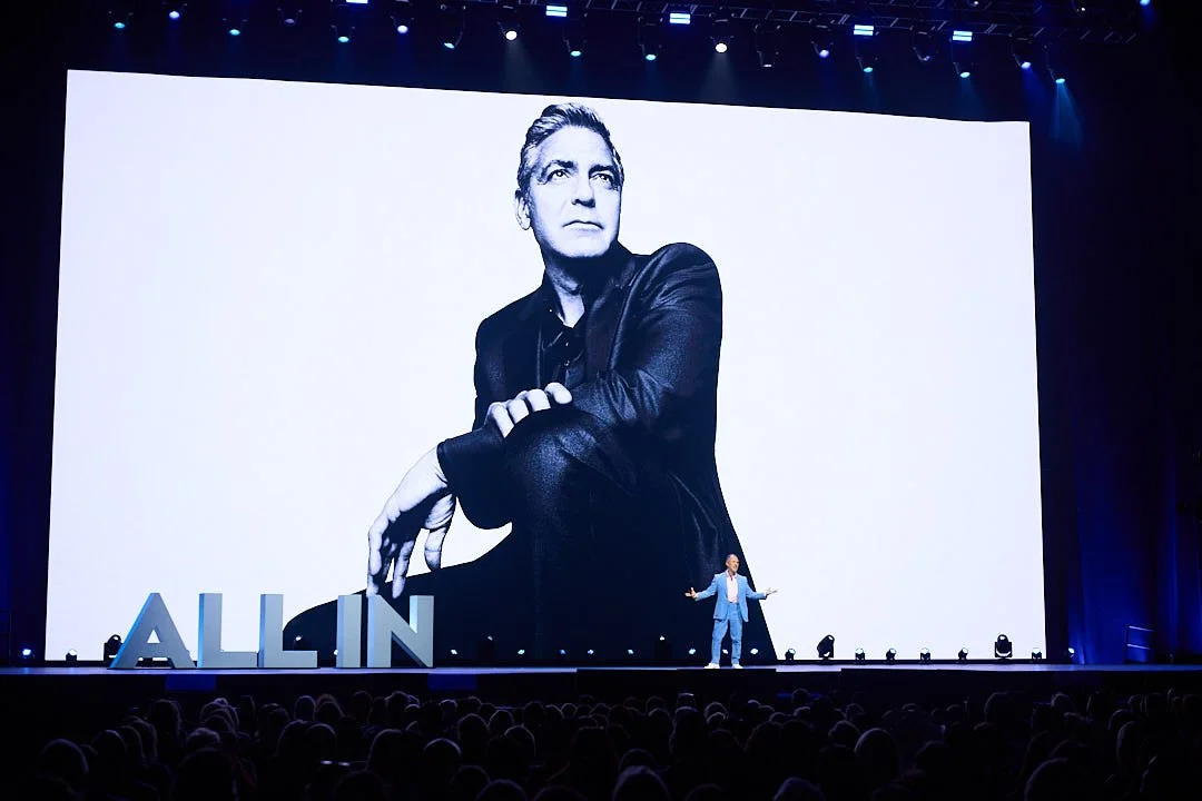 A man in a light blue suit is speaking on stage in front of a large screen displaying a black-and-white photo of himself. The stage has large text that says 'ALL IN'.
