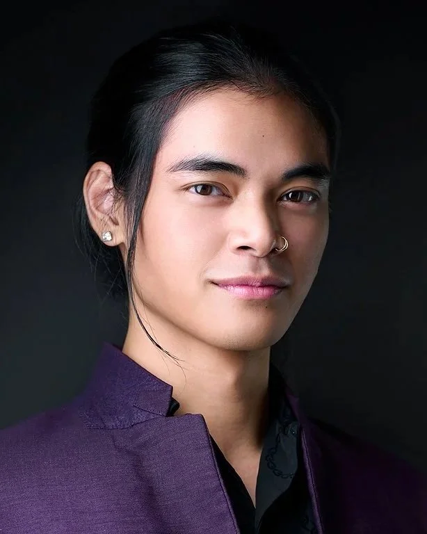 A young person with long dark hair tied back, wearing earrings, and a nose ring. They are dressed in a dark purple jacket and a black shirt, with a confident expression on their face against a dark background.