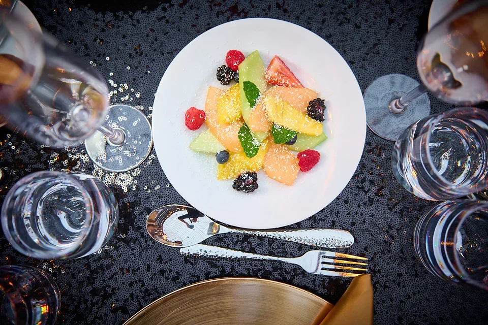 A white plate with mixed fresh fruit slices and berries, set on a black sequin tablecloth, surrounded by four glasses of water, a spoon, and a fork.