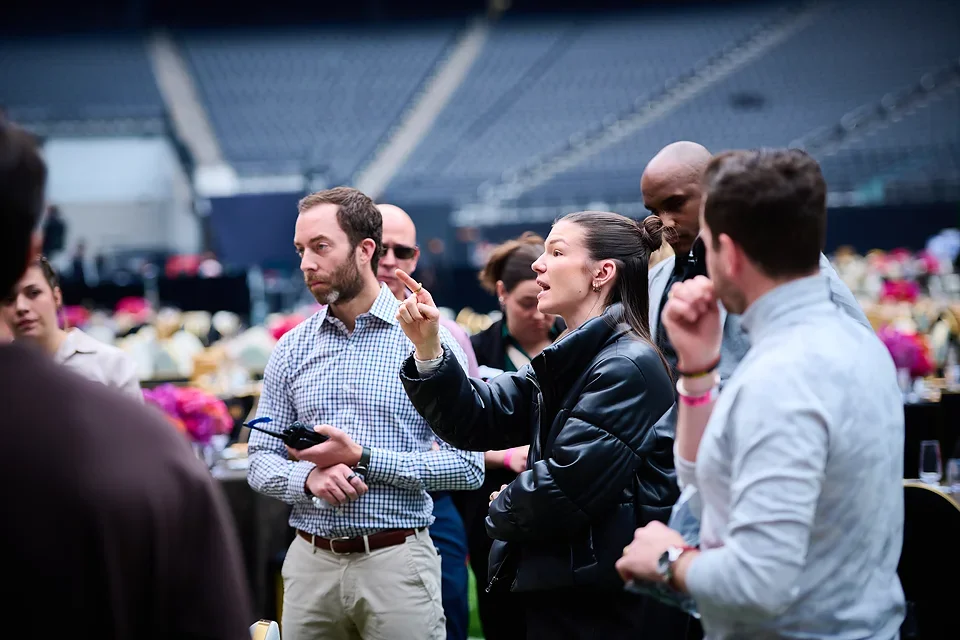 Group of people having a discussion at an indoor event with floral centerpieces on tables.