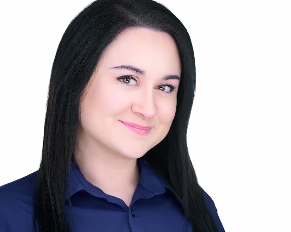 A young woman with black hair and fair skin, wearing a navy blue shirt, smiling lightly with a plain white background.
