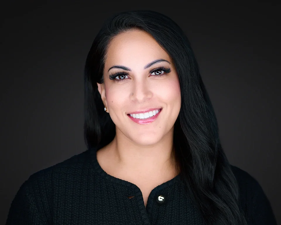 Portrait of a woman with long black hair, smiling, wearing a black top against a dark background.