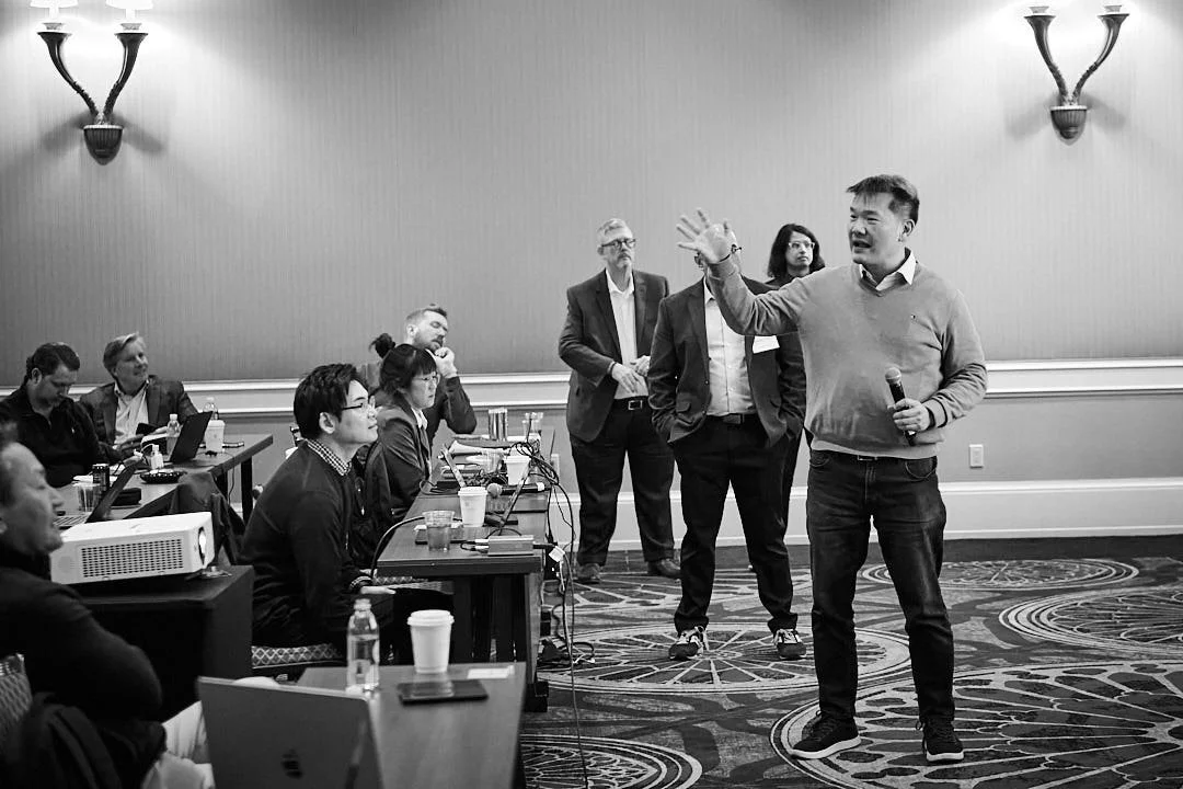 A man holding a microphone and gesturing toward an audience in a conference room with several seated attendees, some with laptops, and a few standing in the background.
