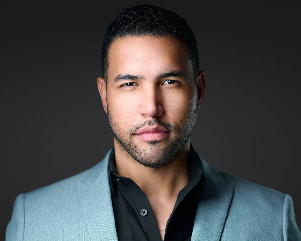 A man with short black hair, a light beard, and light brown skin wearing a light gray blazer over a black shirt, looking directly at the camera with a serious expression against a dark background.
