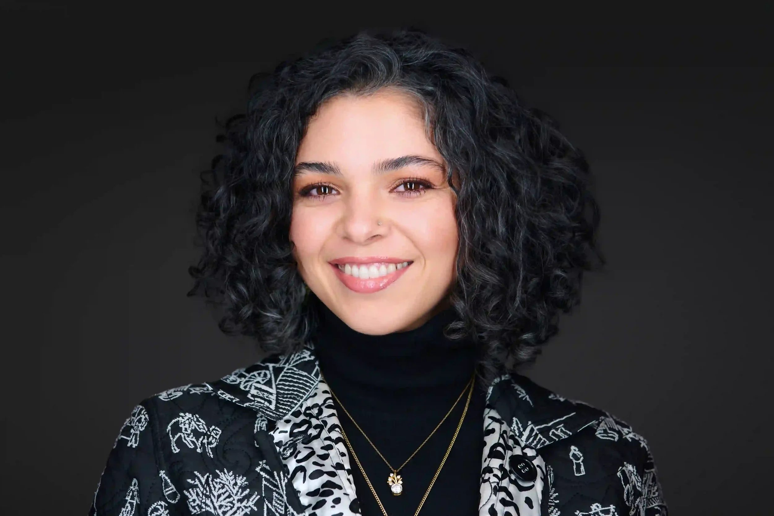 Portrait of a woman with curly black hair, smiling, wearing a black turtleneck, patterned blazer, and layered gold necklaces.
